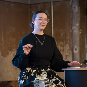 Senior architect Stephanie Kyle speaking at a conference about neurodiversity and inclusive design, gesturing while seated at a desk.