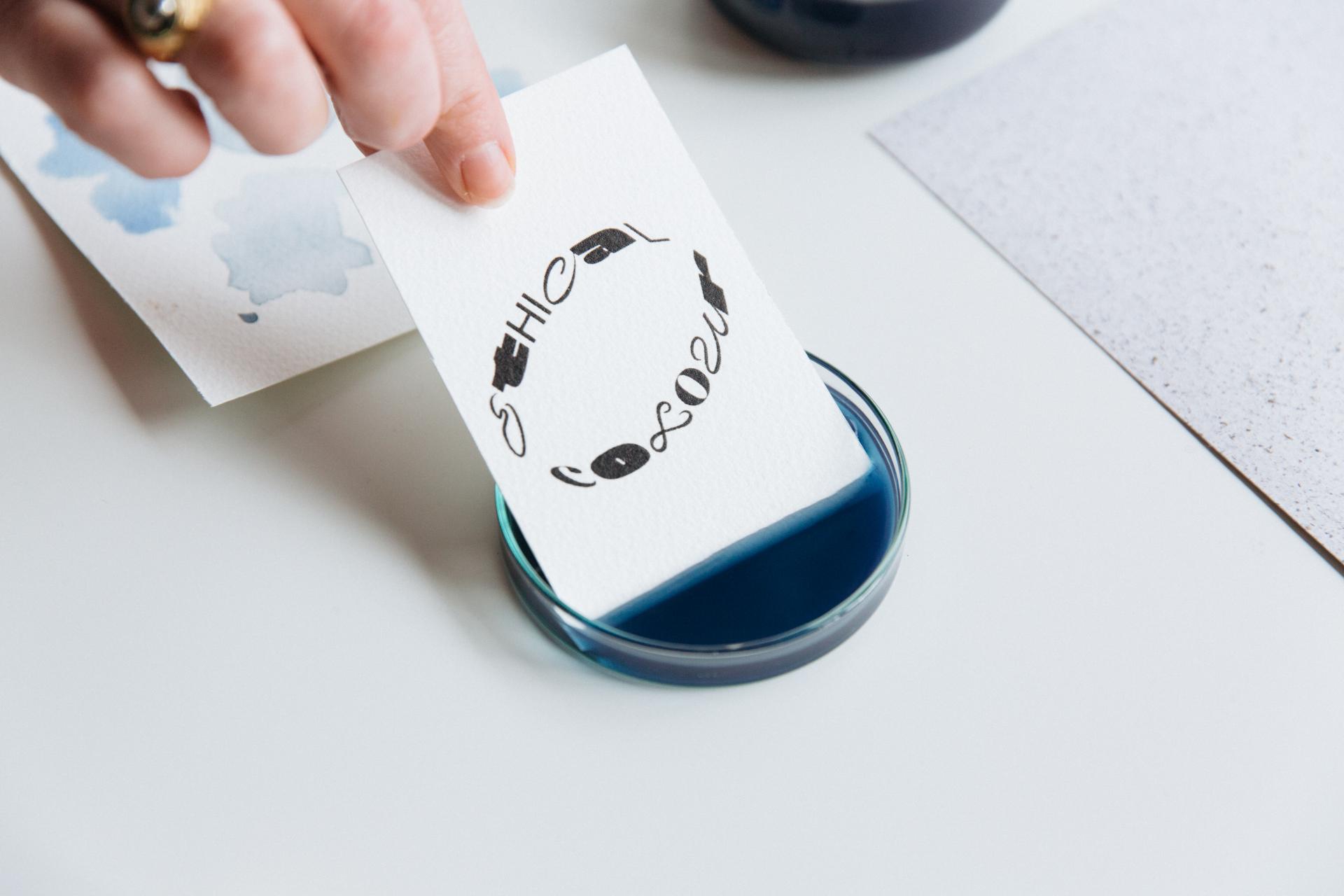 A hand immerses a paper with "Ethical Colour" text into a blue dye, representing sustainable color practices in design.