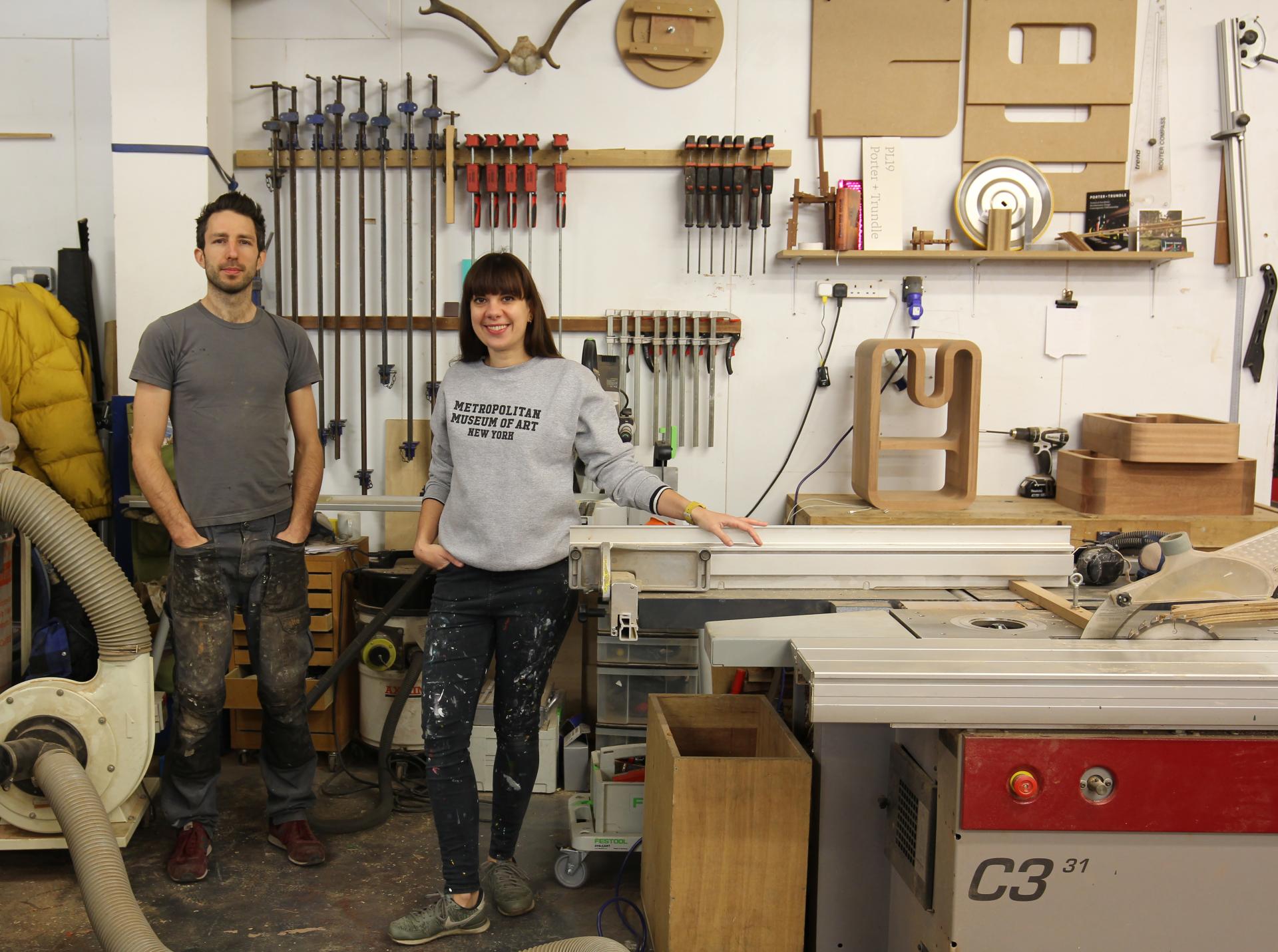 Two designer-makers stand in a workshop filled with woodworking tools, showcasing the craftsmanship and creativity of Design-Nation.
