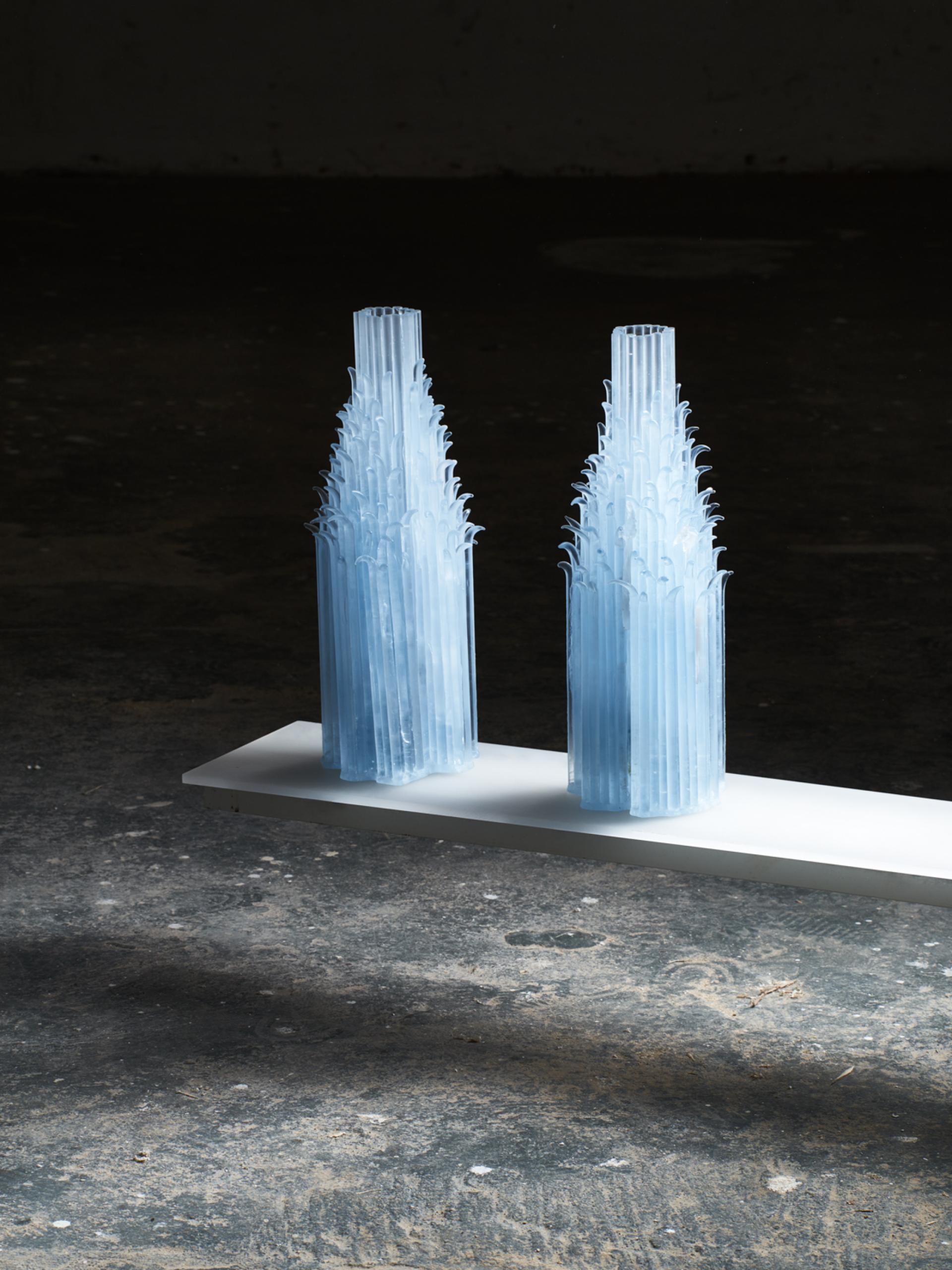 Two elegant blue glass sculptures, resembling tall bottles, reflecting light in a dark exhibition space.
