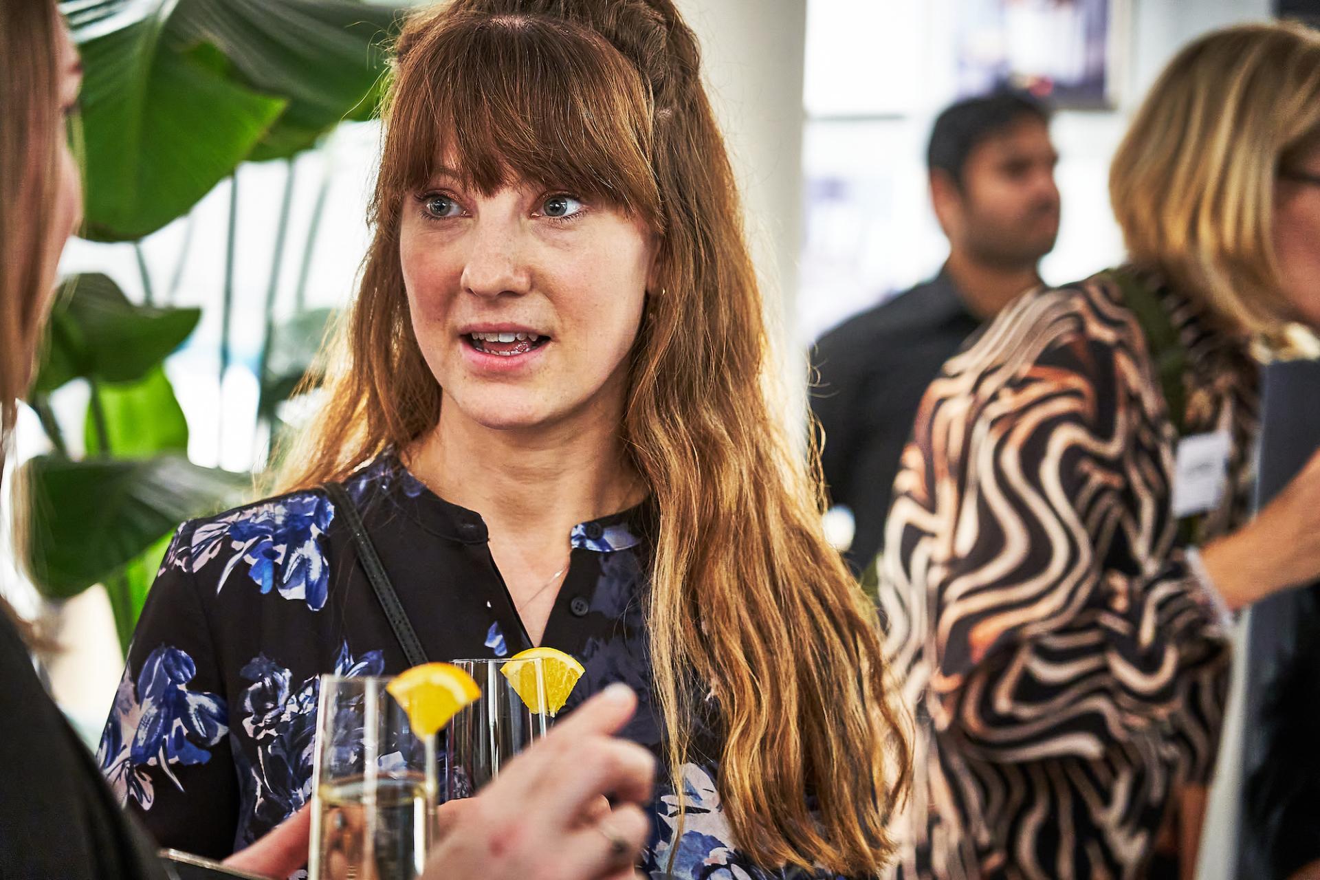Engaged woman at a networking event, discussing the podcast 42 Roles in the Built Environment while holding cocktails.