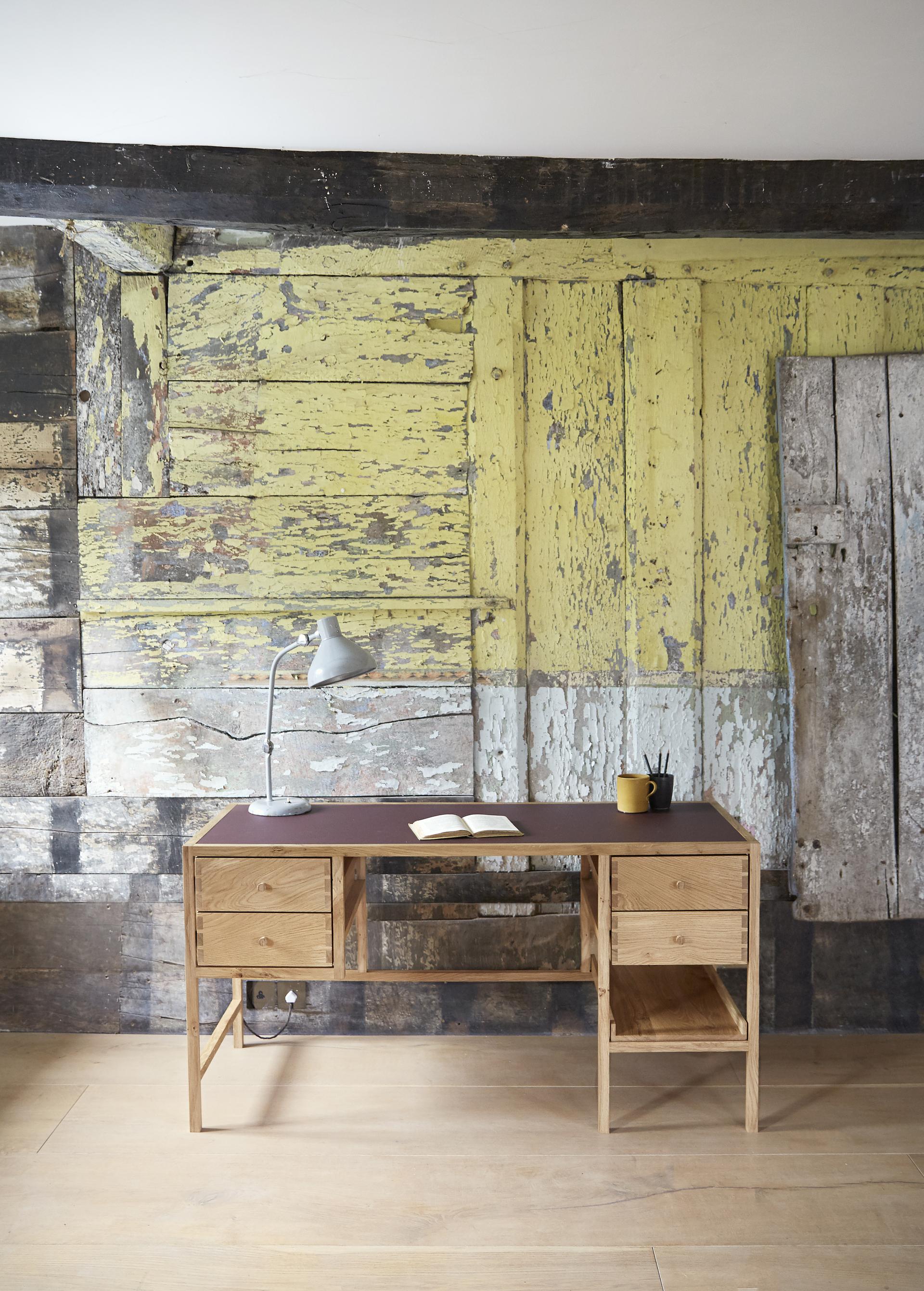 Stylish wooden desk with a lamp, notebook, and yellow weathered wall backdrop, showcasing contemporary design.