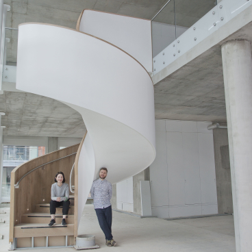 Designers Rebecca May and Michael Wild pose beside a modern spiral staircase in an industrial-style studio space.