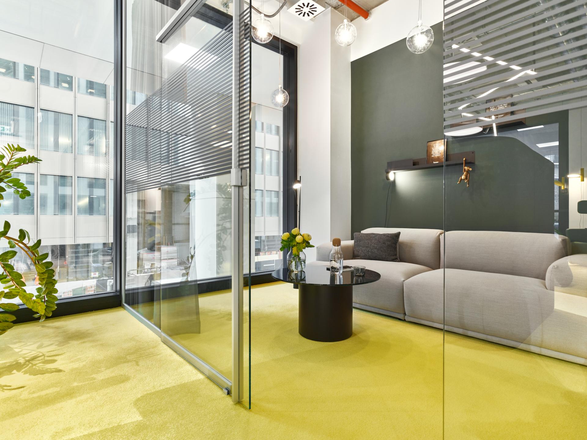 Modern office space featuring a cozy lounge area with plants, bright yellow carpet, and glass walls, reflecting pandemic-inspired design changes.
