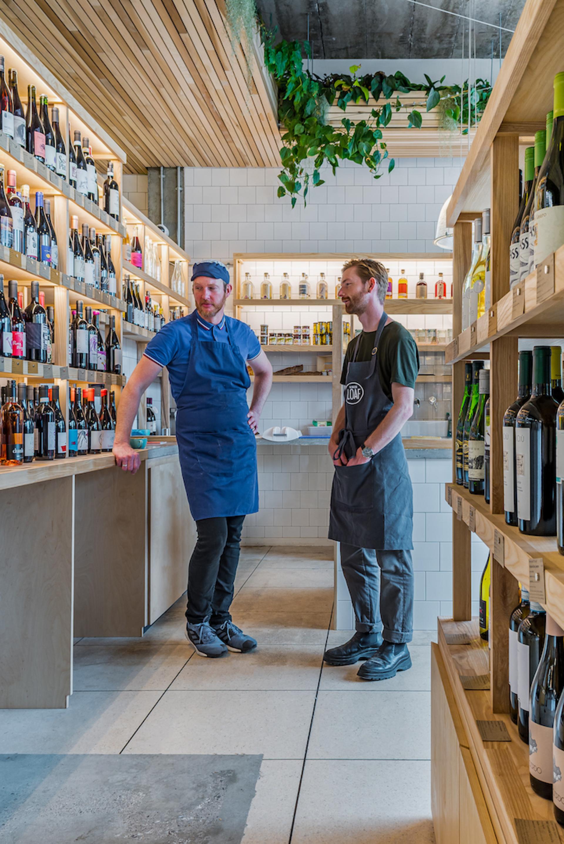 Two men in aprons converse in a stylish interior filled with wine bottles, showcasing The Bristol Loaf’s new design.