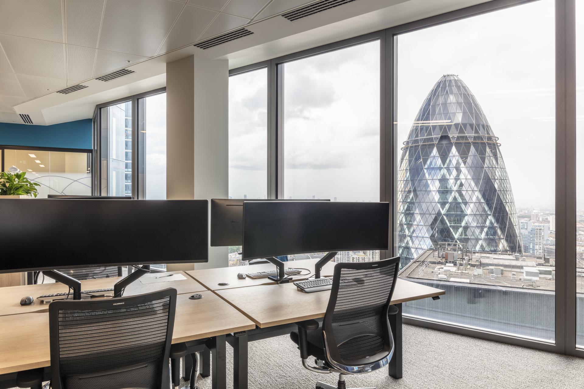 Modern office space with dual monitors, featuring views of London's skyline and the iconic Gherkin building.