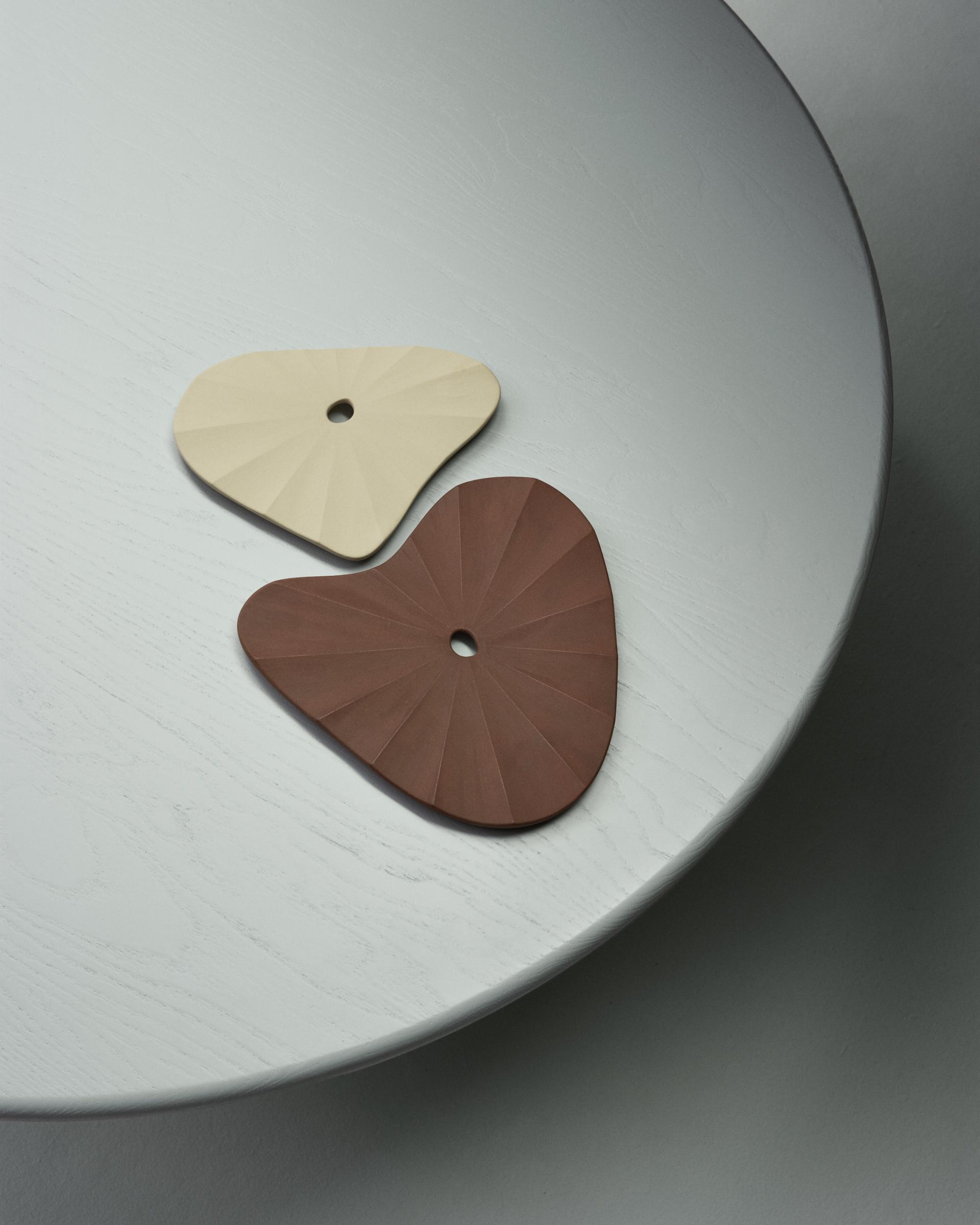 Two uniquely shaped, textured ceramic coasters in beige and brown placed on a round white tabletop, highlighting circular design.