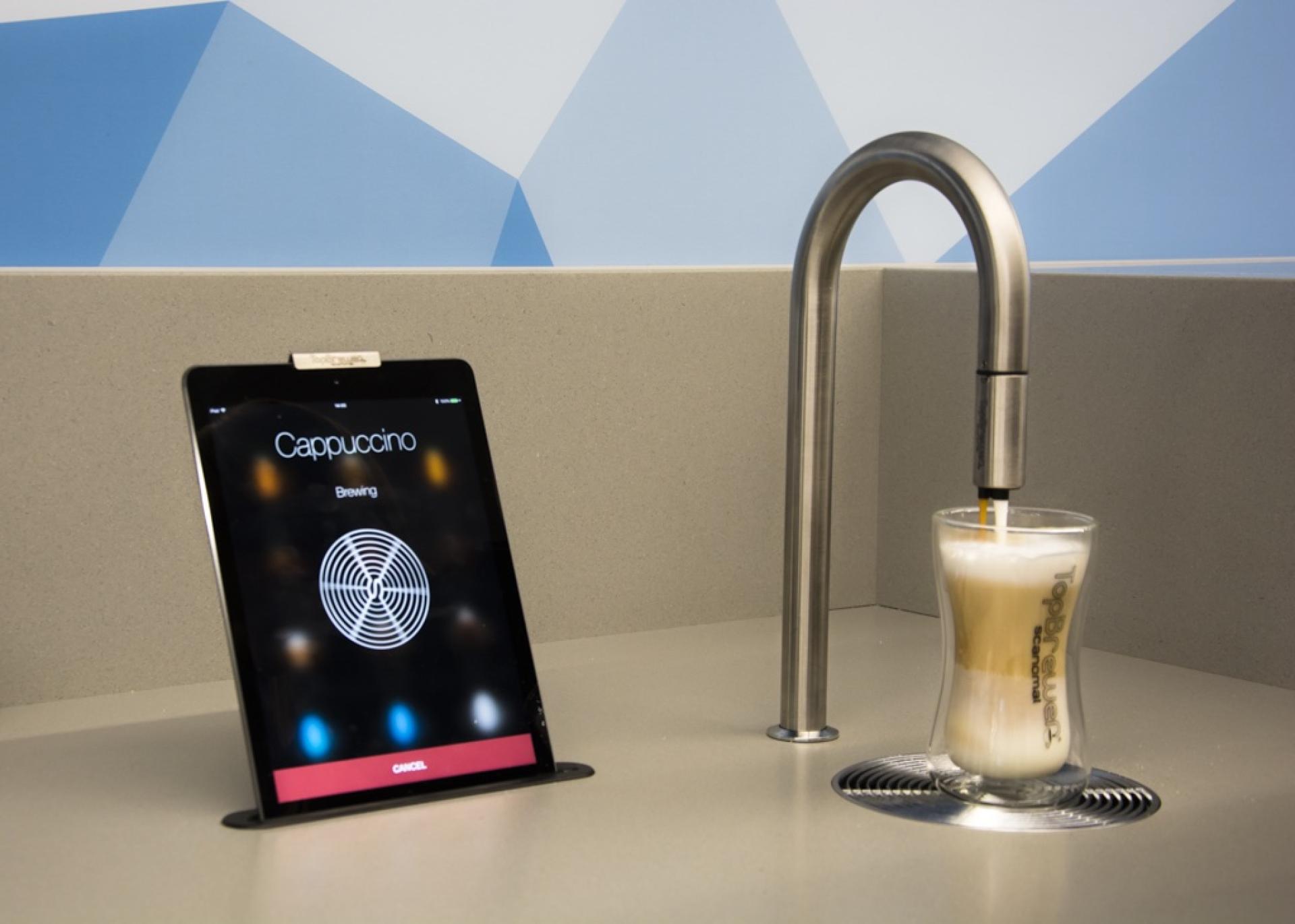 Cappuccino brewing on a tablet next to a glass, showcasing Scanomat's TopBrewer coffee solution for modern workplaces.
