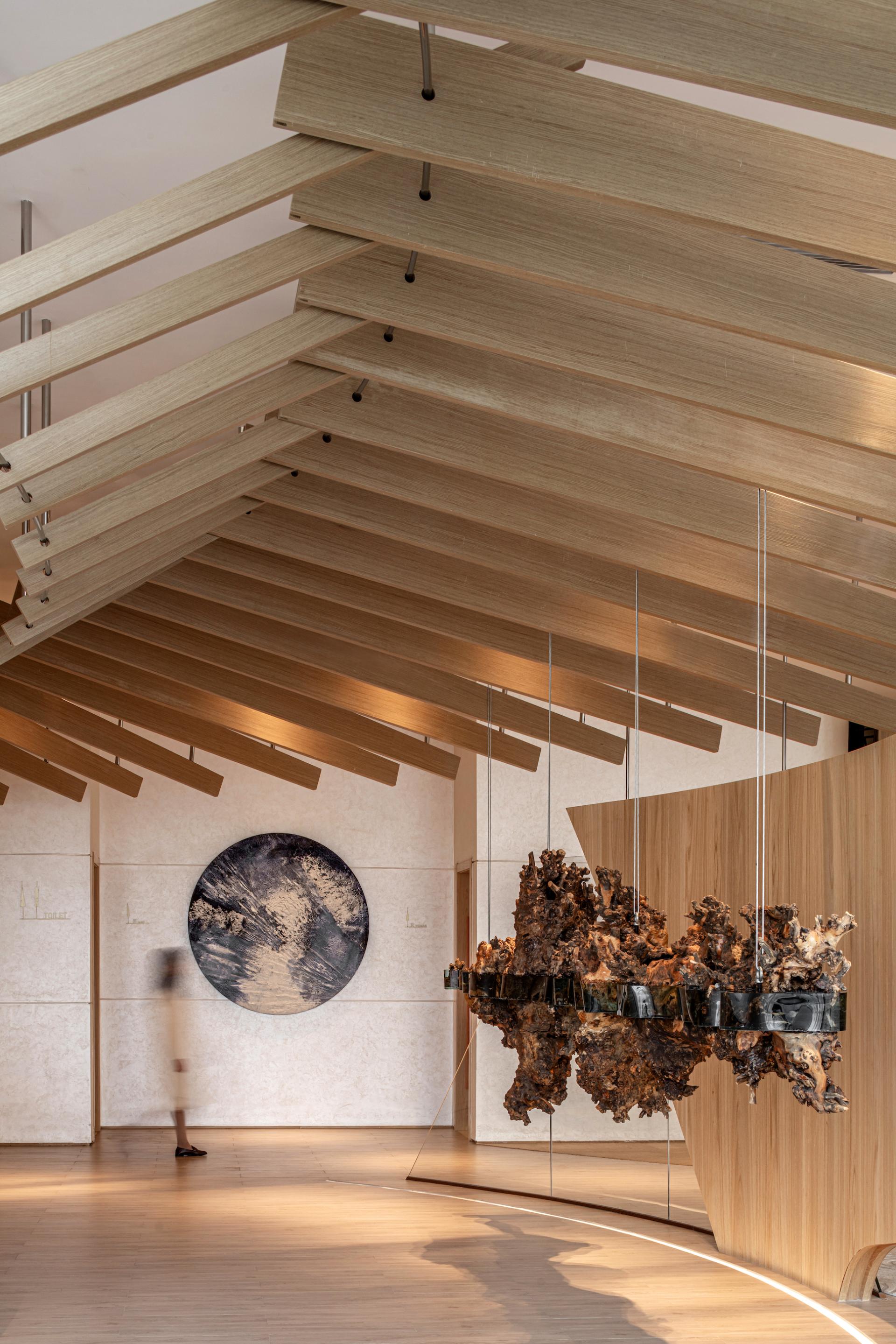 Sculptural wooden ceiling beams highlight an elegant exhibition hall featuring contemporary art installations in lush greenery.