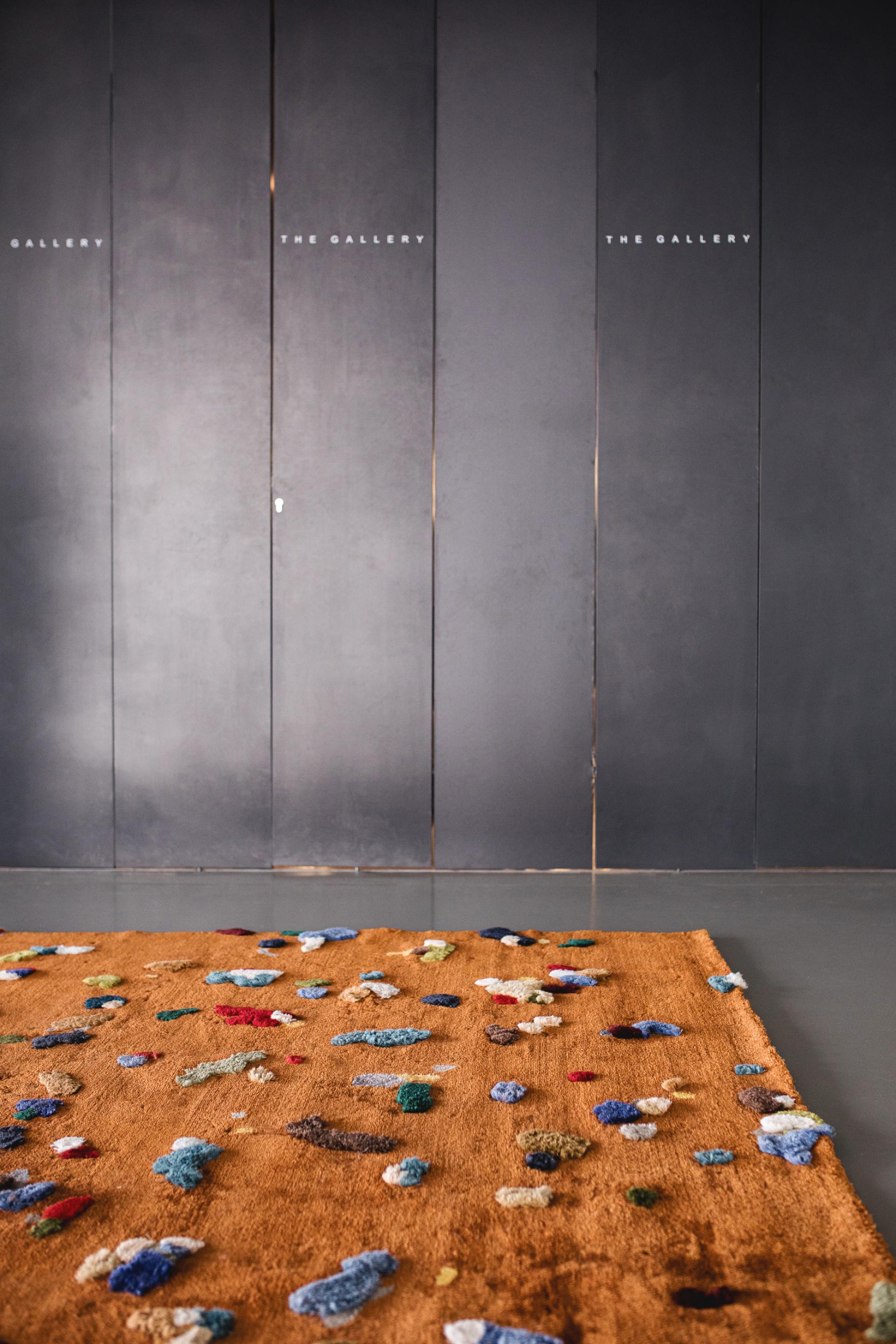 Chaos Rug by Audronė Drungilaitė featuring vibrant multicolored patches on a warm-toned background in a gallery setting.