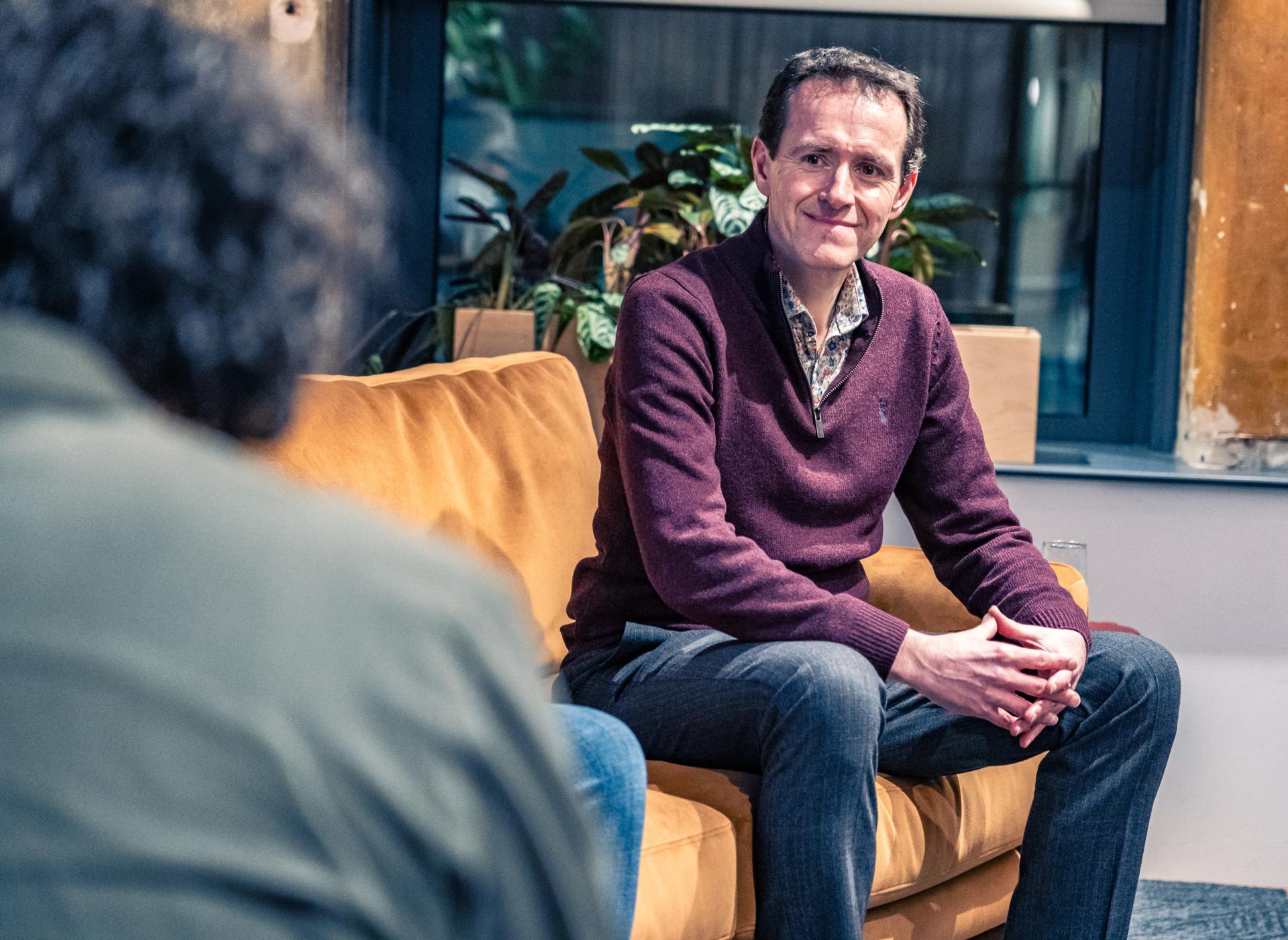 Discussion on achieving Net Zero, featuring a man sitting on a couch with plants in the background.