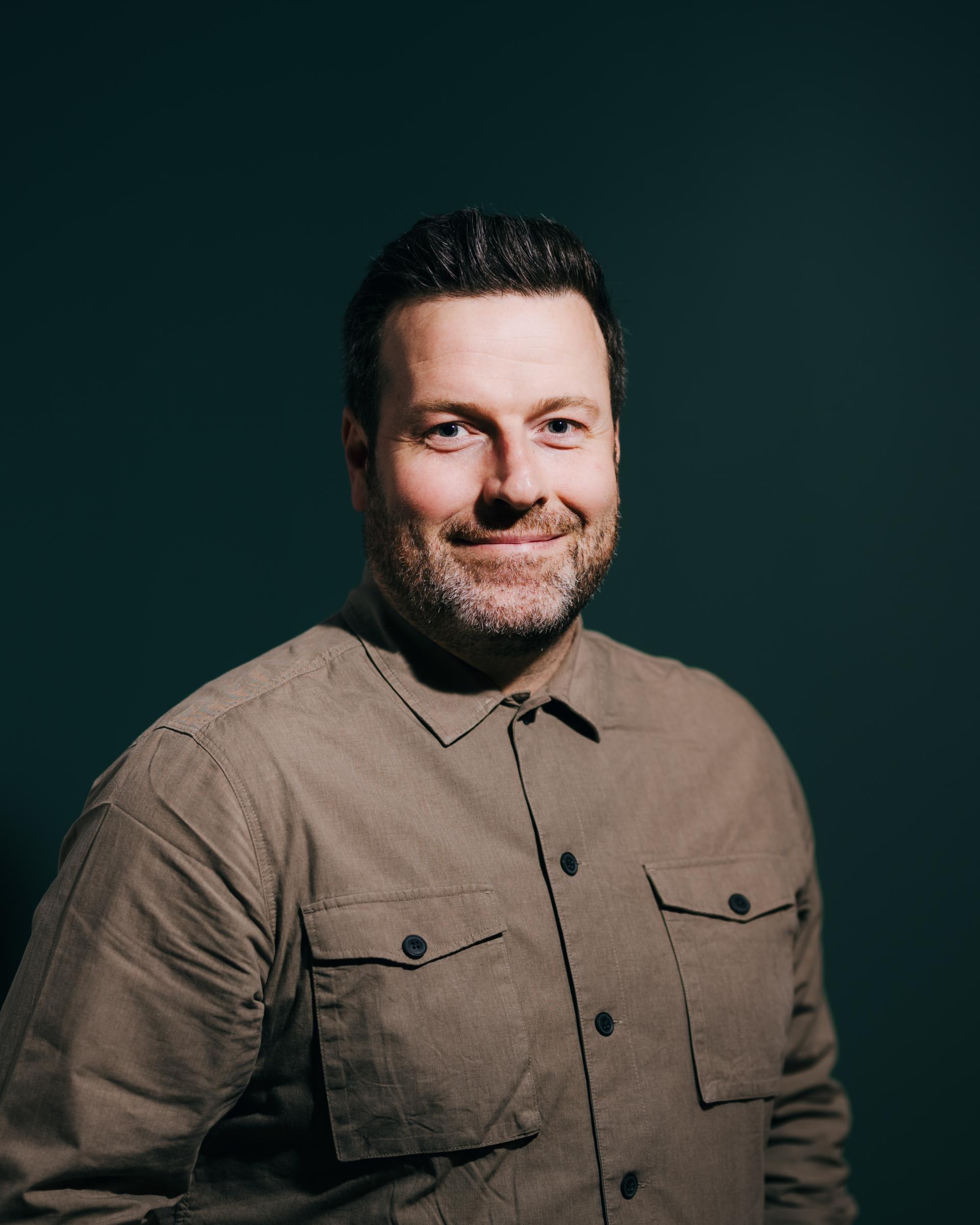 Man with a beard and brown shirt smiles against a dark green background, representing neurodiversity discussion.
