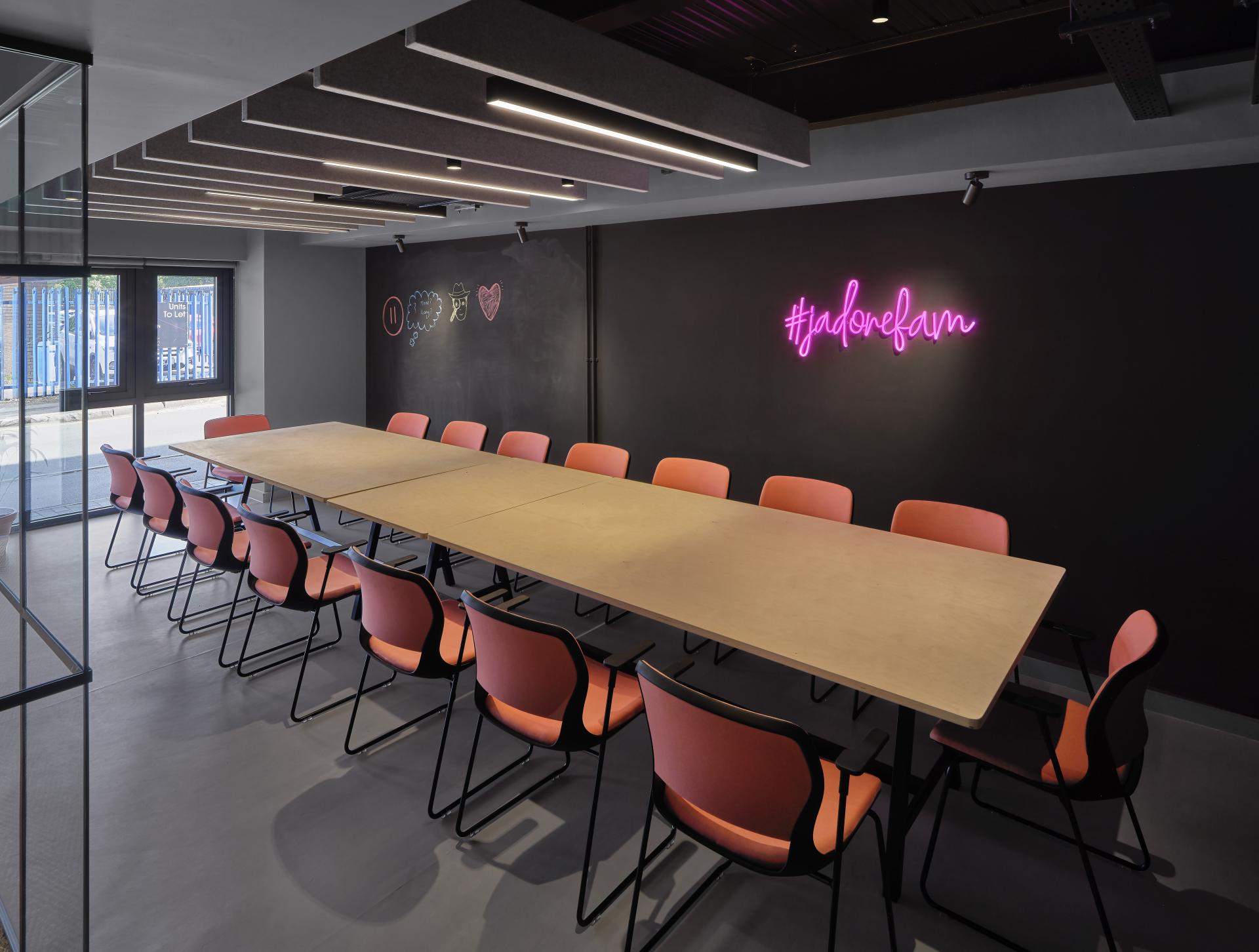 Modern conference room featuring a long table, pink chairs, and neon "#jadorefam" sign on the wall.