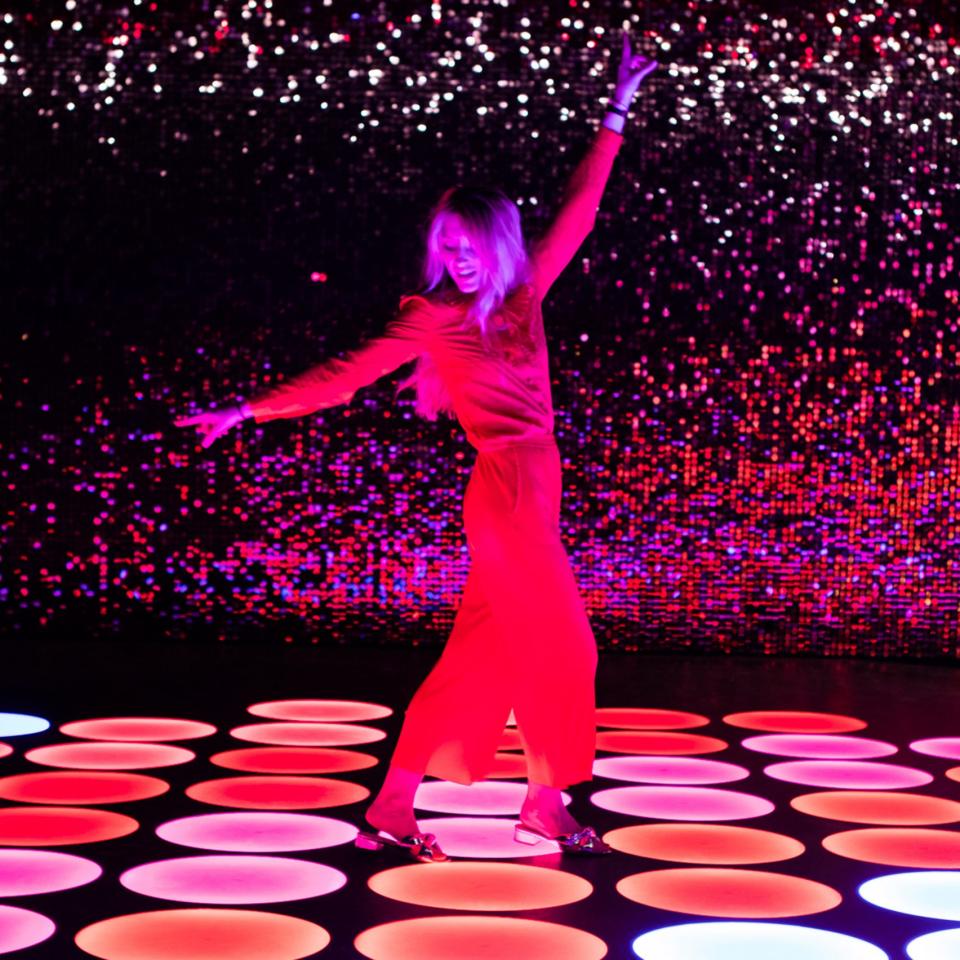 A woman dances joyfully on colorful illuminated platforms in the vibrant Color Factory exhibition in New York City.