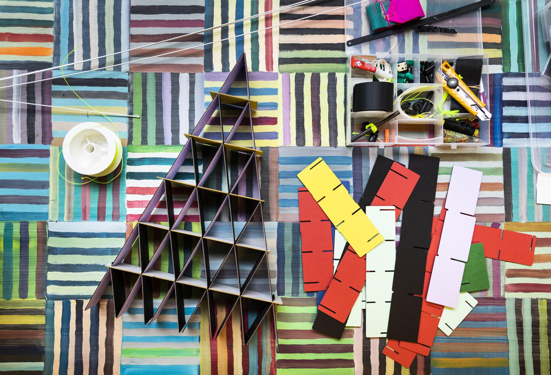 Colorful materials and tools scattered on a striped surface, featuring a geometric paper structure, embodying Hem's design creativity.