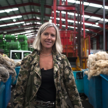 Designer Ella Doran stands amid bales of British wool in a textile warehouse, showcasing the wool's transformative journey.