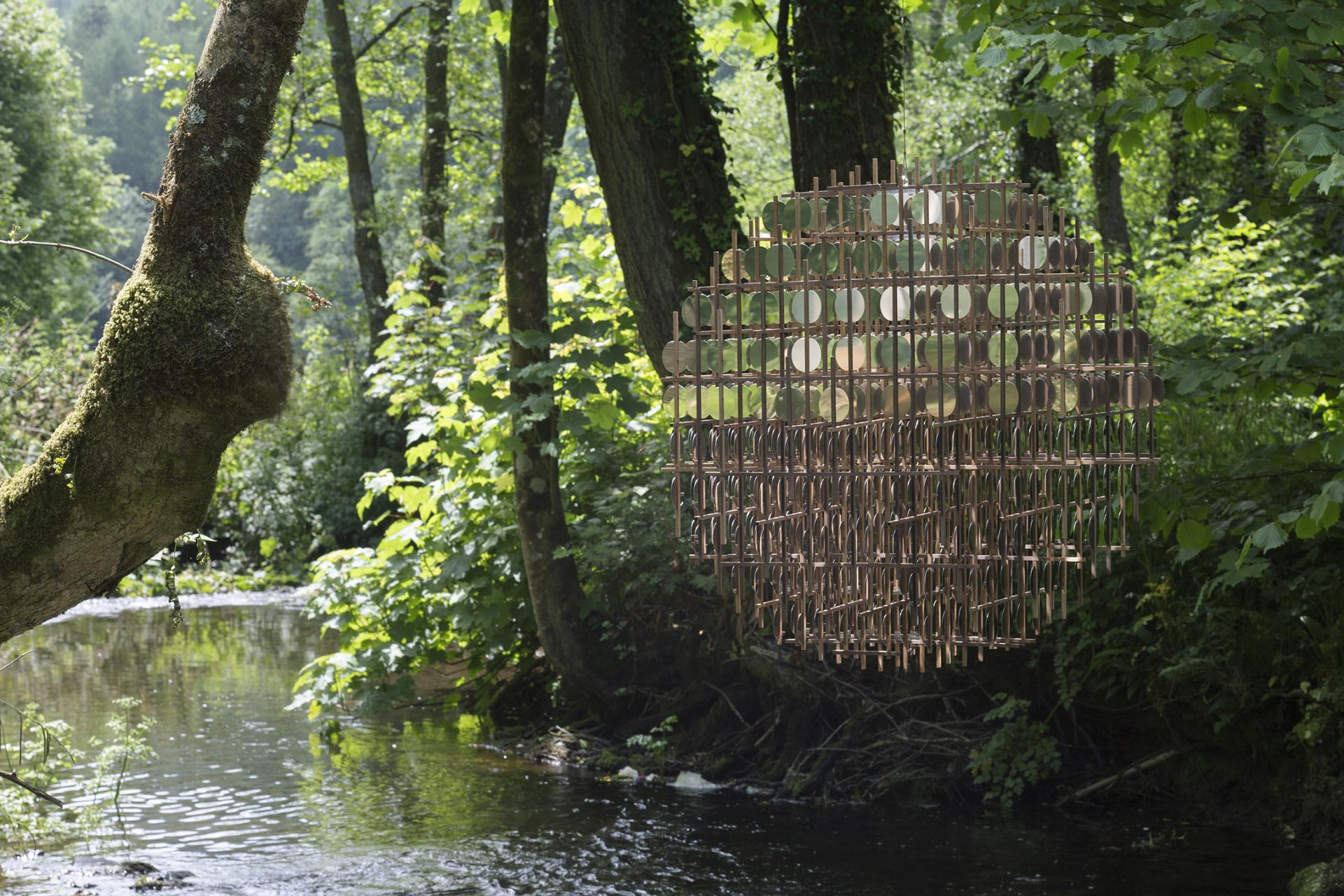 Sculptural installation by Giles Miller suspended in a lush green forest by a serene stream, reflecting natural light.