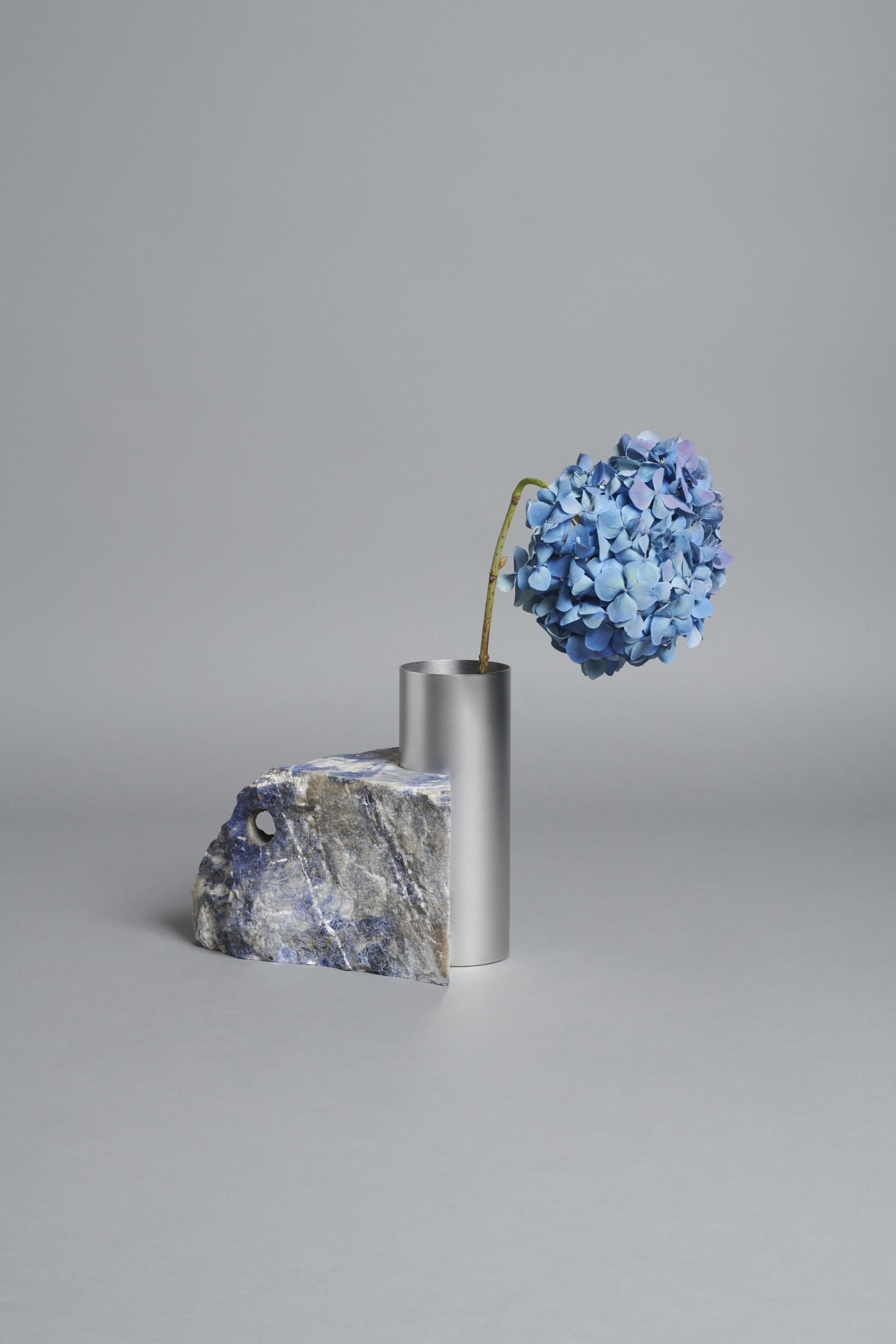 Stylish vase made of metal holds blue hydrangeas, complemented by a textured stone, showcasing raw design aesthetics.