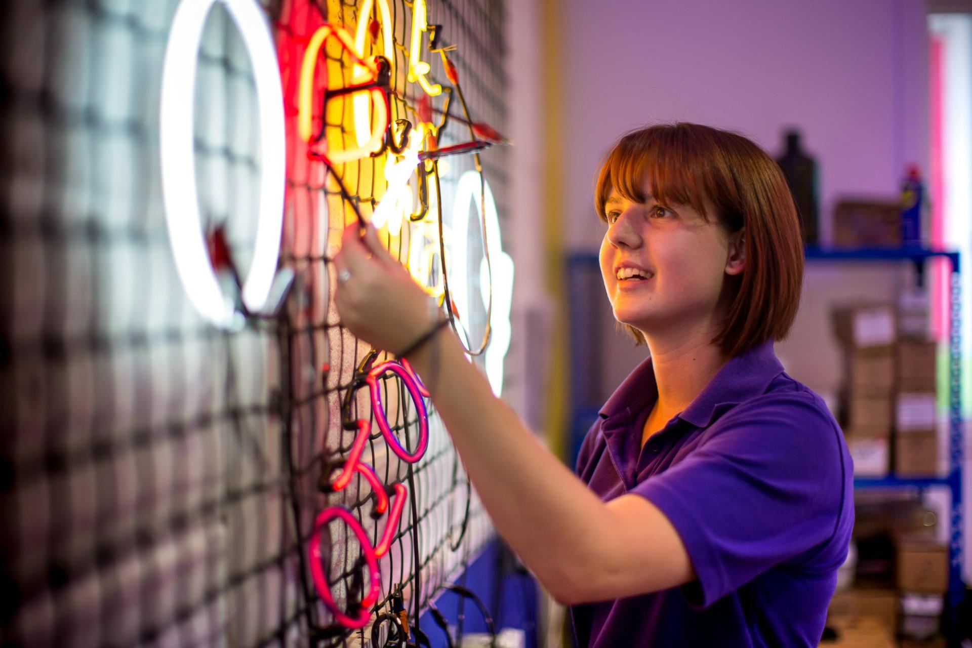 A woman adjusts colorful neon signs, showcasing craftsmanship and creativity in the manufacture of vibrant illuminated displays.