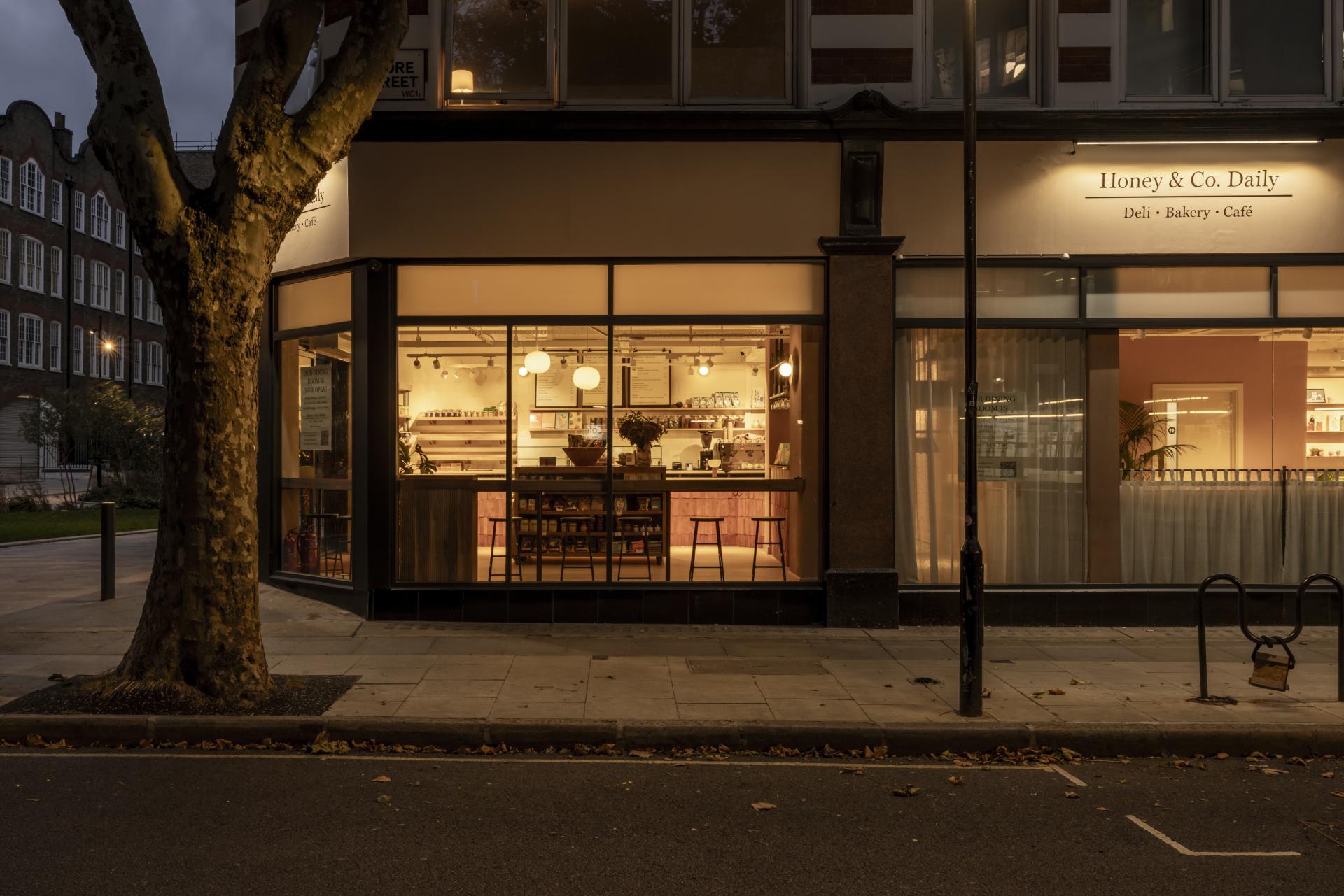 Honey & Co Daily deli and café illuminated at night, showcasing a warm interior with terracotta accents and inviting ambiance.