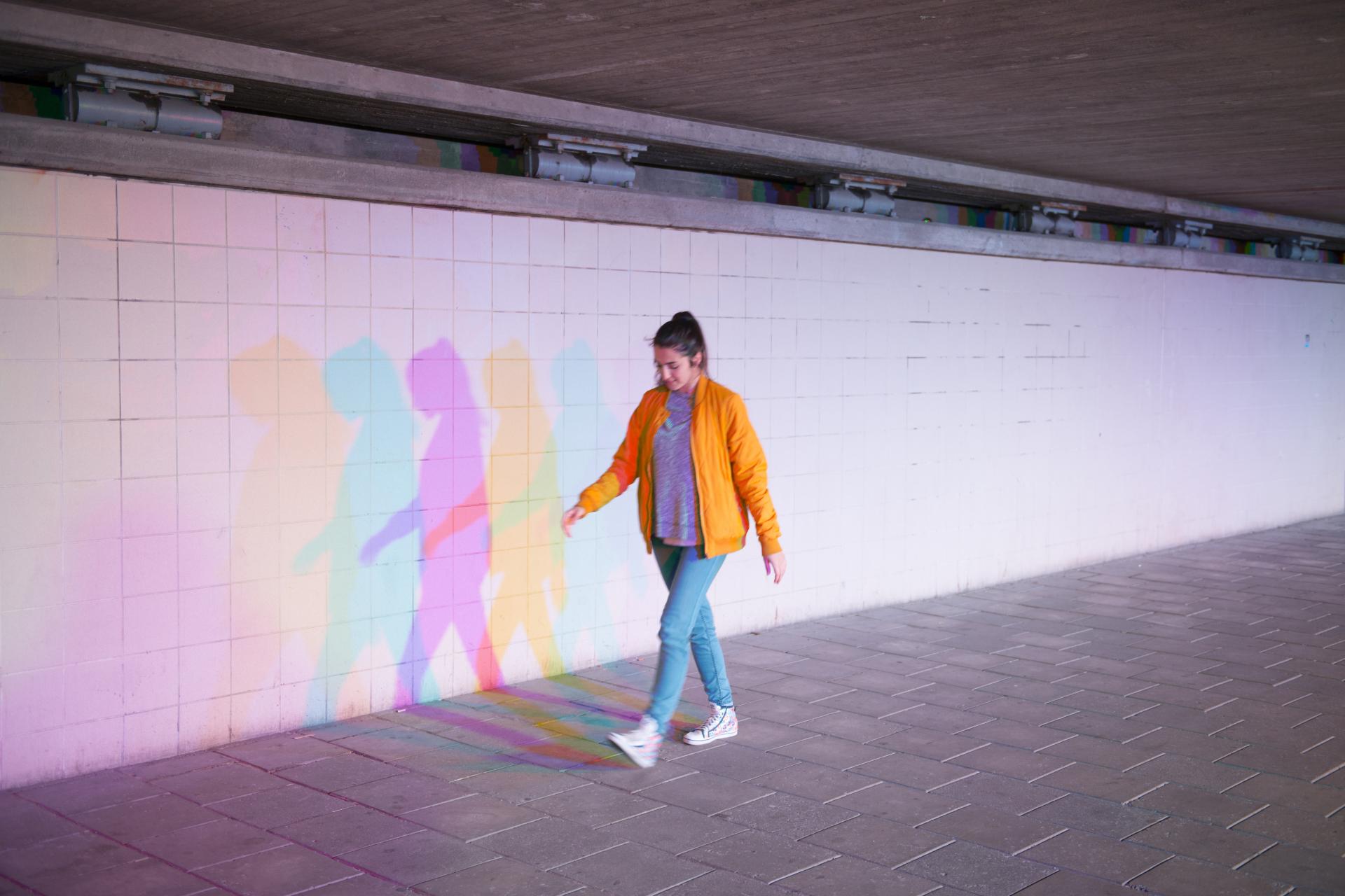 A woman in an orange jacket walks past a colorful shadow on a wall in an urban setting.