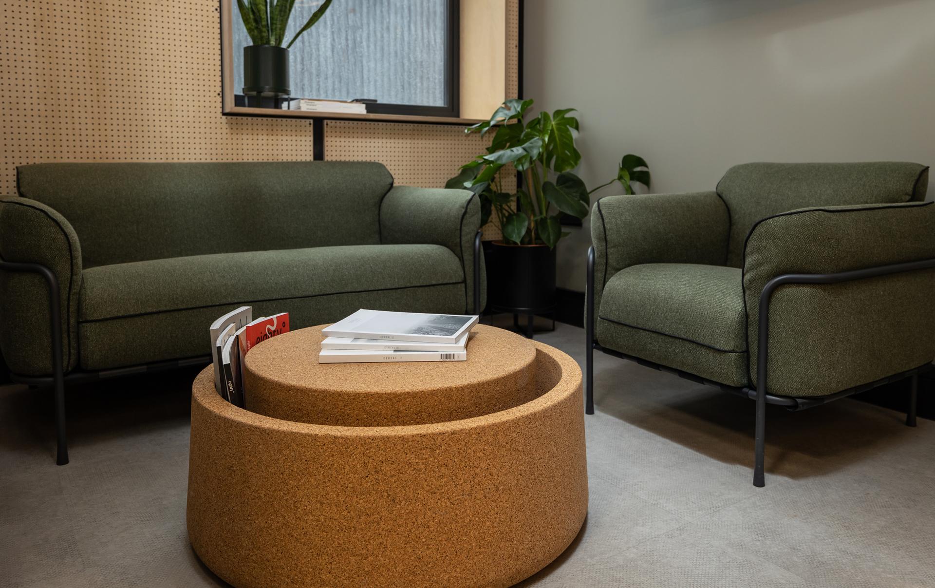 Modern lounge area featuring green sofas, a cork coffee table, and indoor plants in a stylish office space.