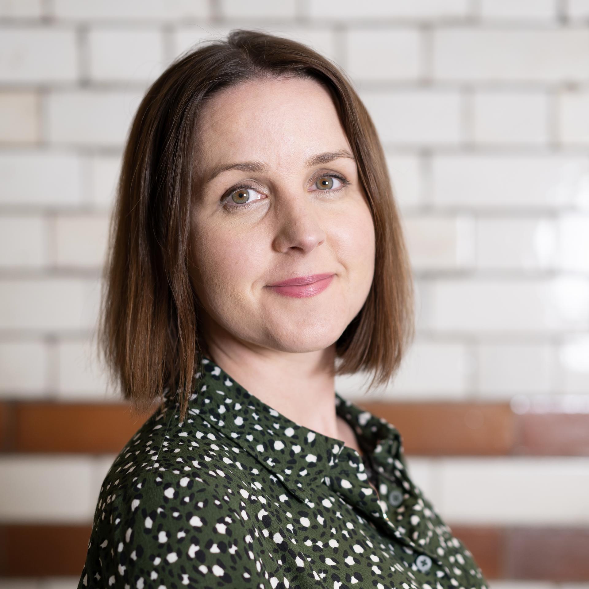 Professional woman with short hair, wearing a patterned green blouse, stands against a modern brick wall.