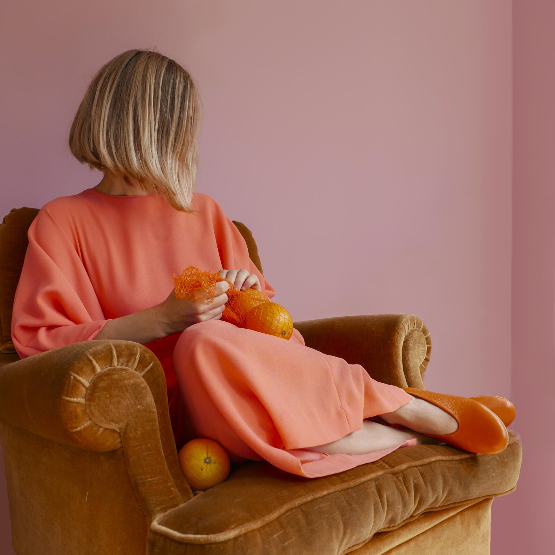 A woman in a coral outfit seated on a golden chair, playfully holding oranges against a pink backdrop.