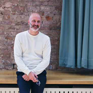 Jasper Sanders, designer and founder of Jasper Sanders + Partners, smiling while seated against a brick wall backdrop.