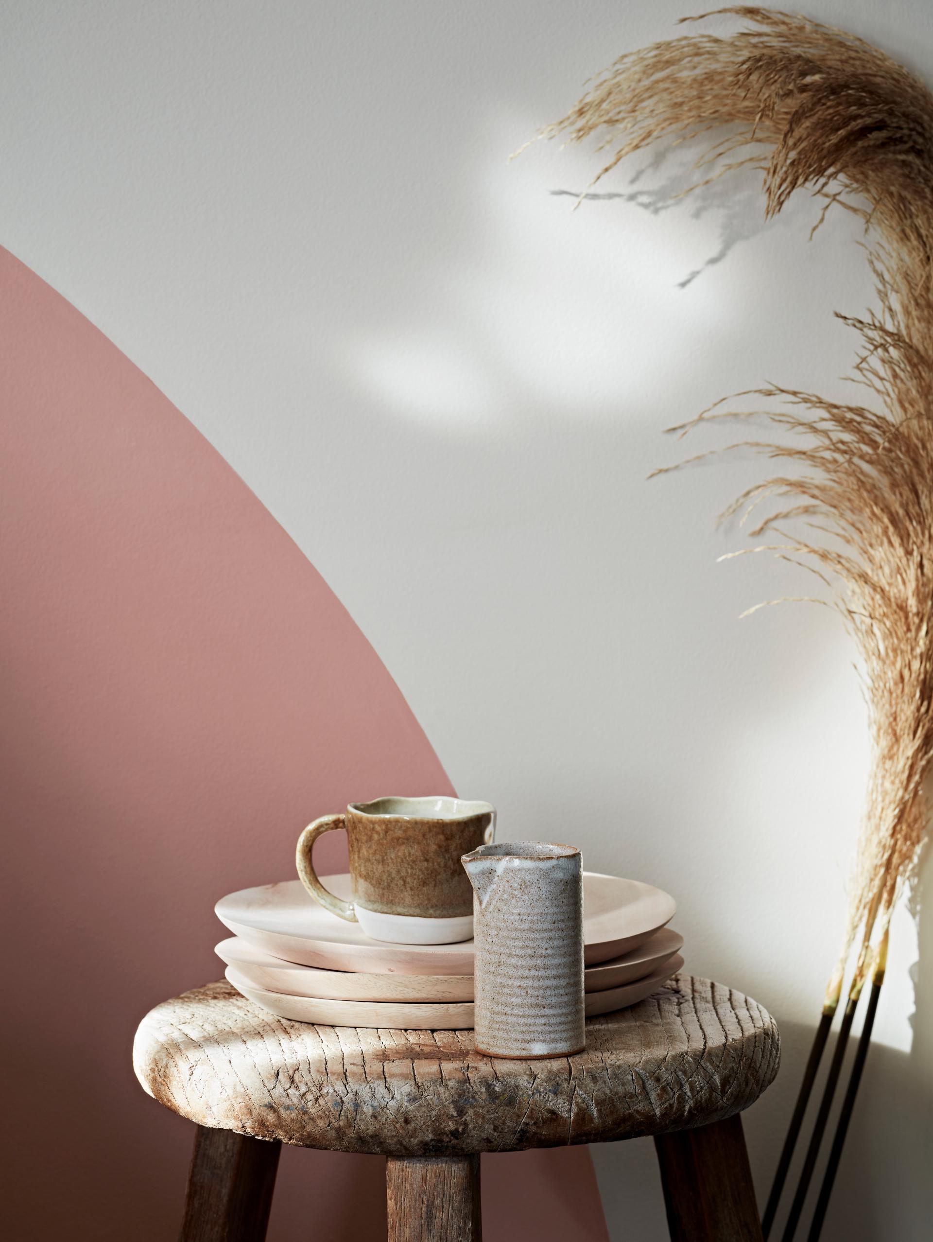 Rustic wooden stool holding handmade ceramic tableware, featuring earthy tones, alongside decorative dried grass and a colorful wall.