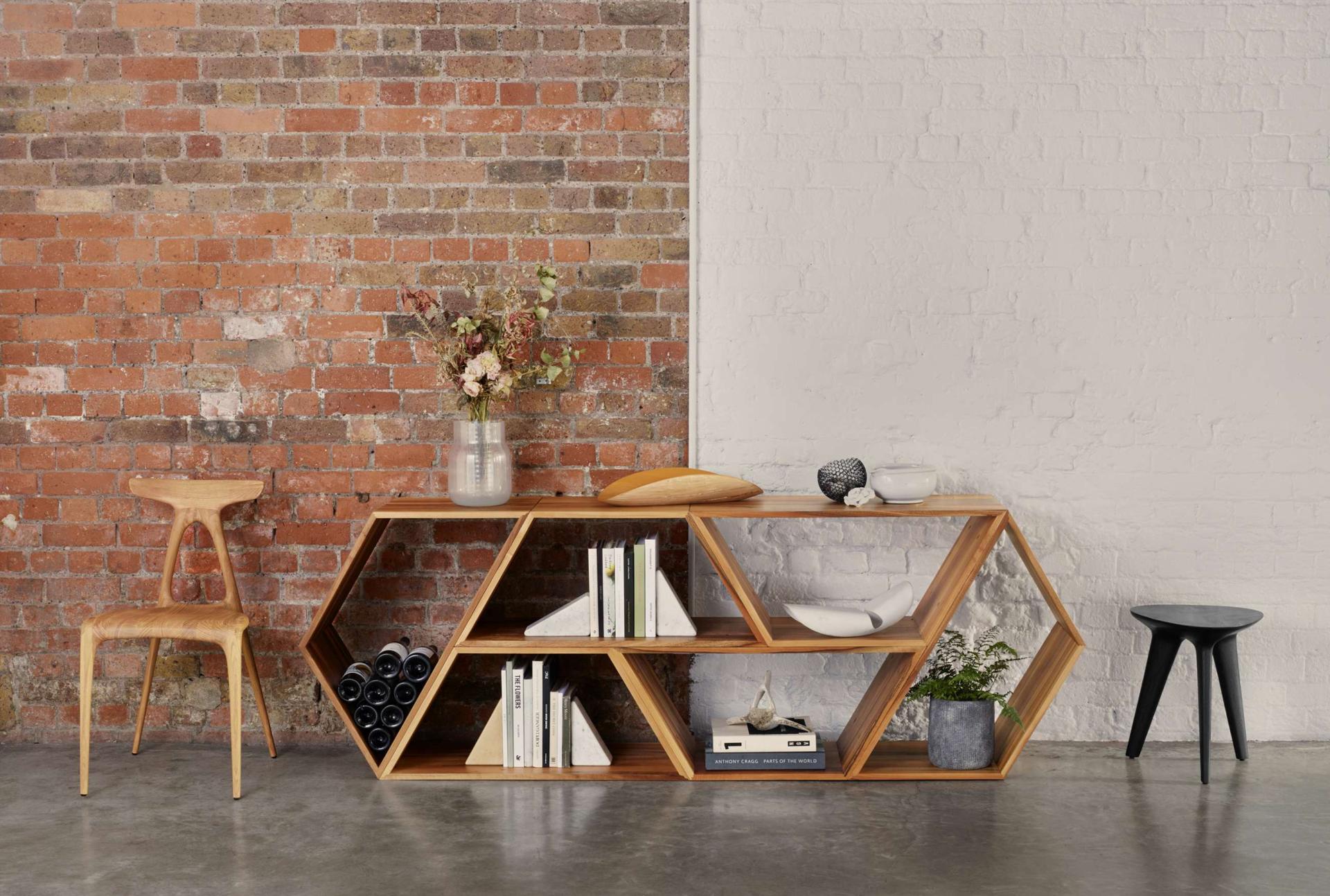 Stylish geometric bookshelf with decorative items and a wooden chair against a rustic brick wall.