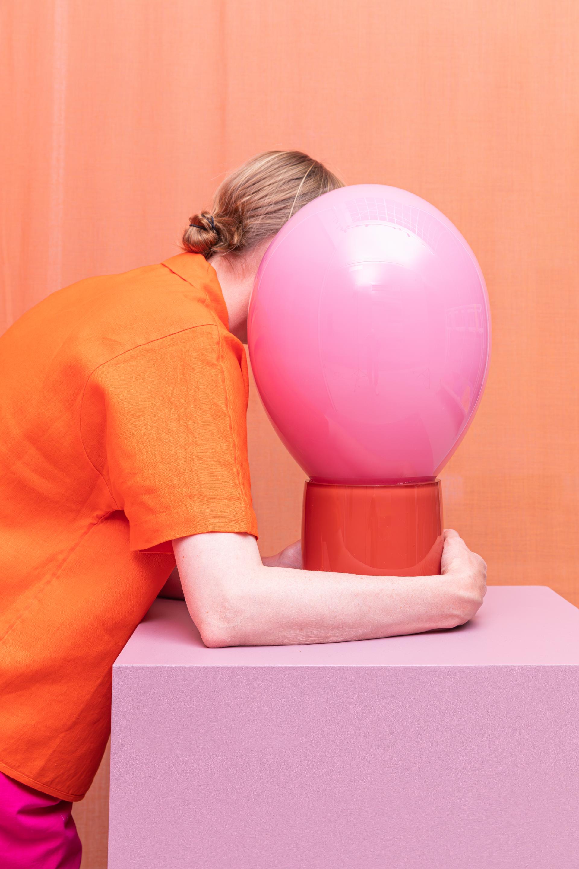 A person embraces a playful pink lamp on a pastel surface, surrounded by vibrant colors highlighting innovative material design.