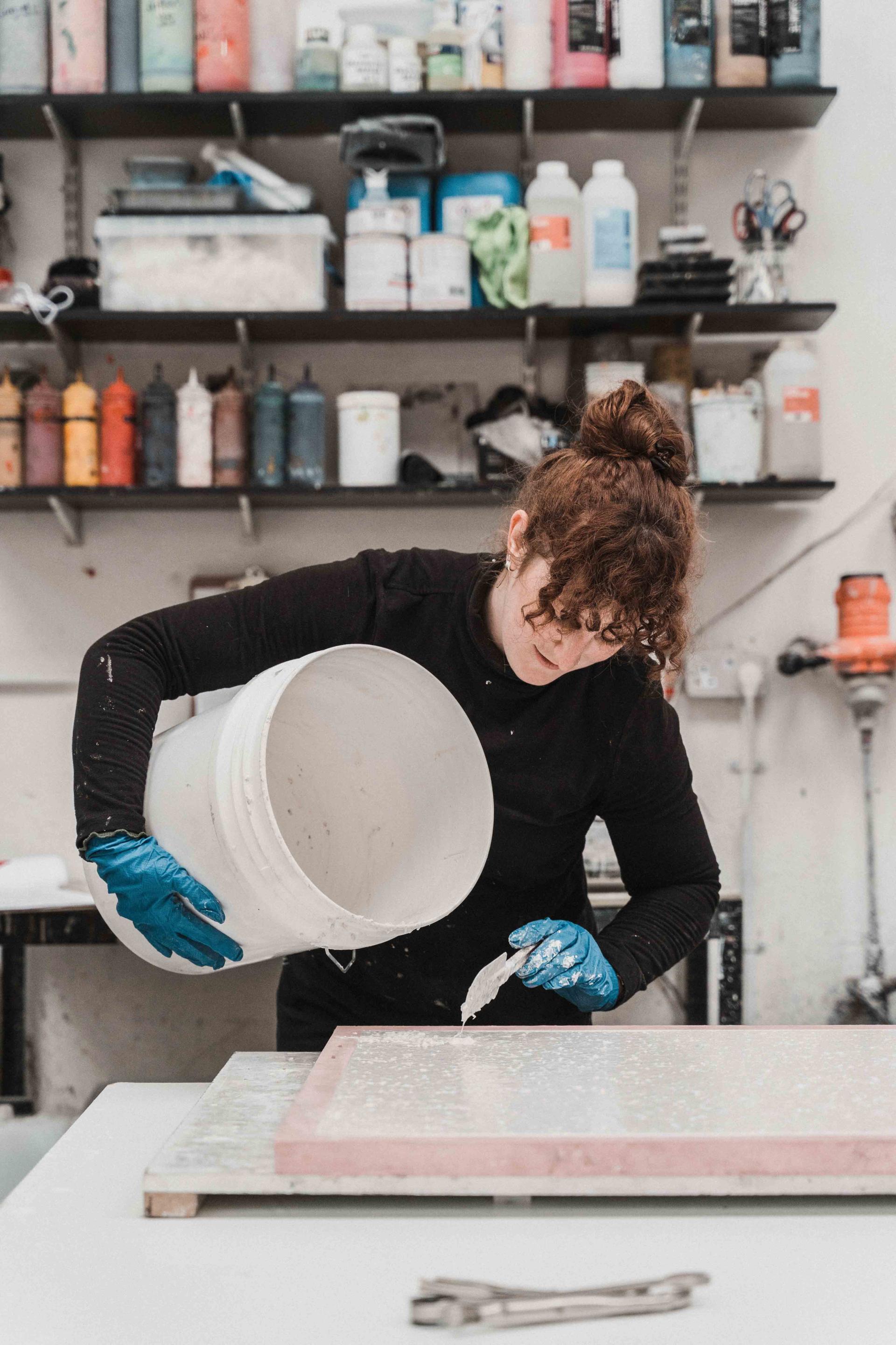 A woman in a black outfit pours material onto a pink ebonite terrazzo sheet in a creative studio.