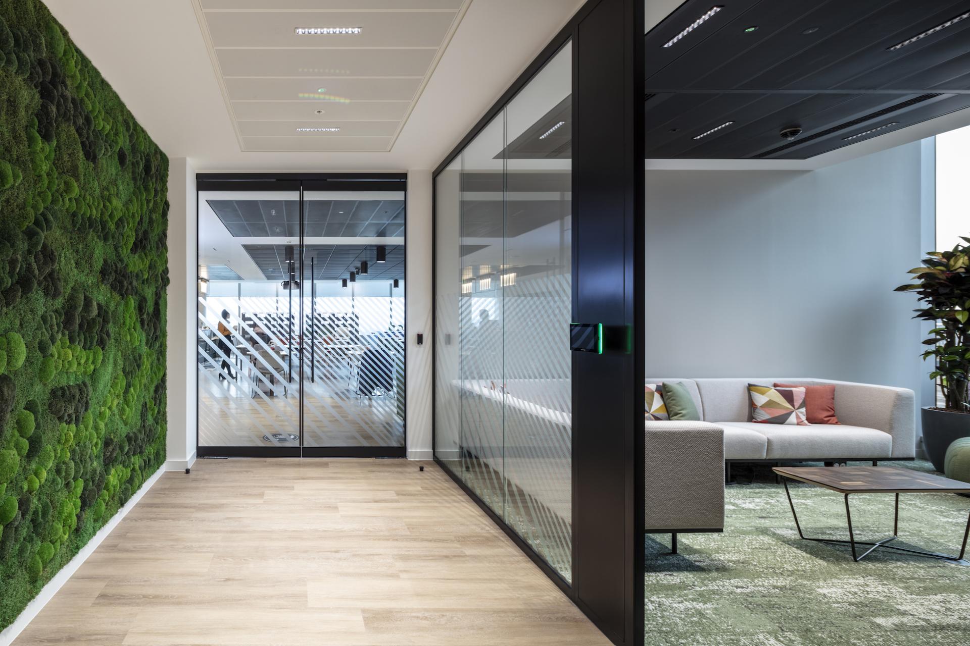 Modern office interior featuring green wall, glass doors, and a cozy seating area, emphasizing contemporary workplace design.