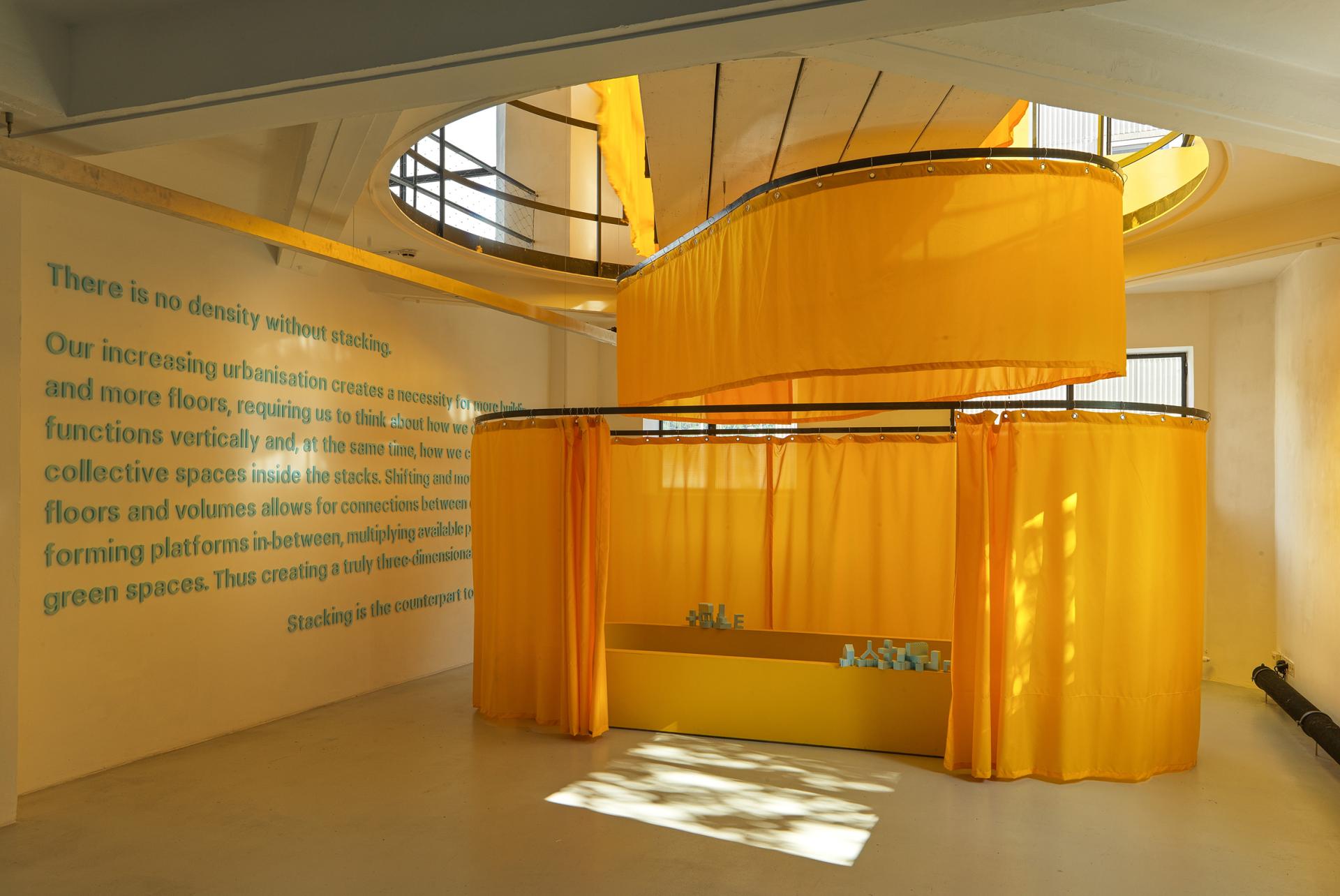 Installation by MVRDV featuring vibrant yellow curtains and architectural models in a spacious exhibition area celebrating urban design.
