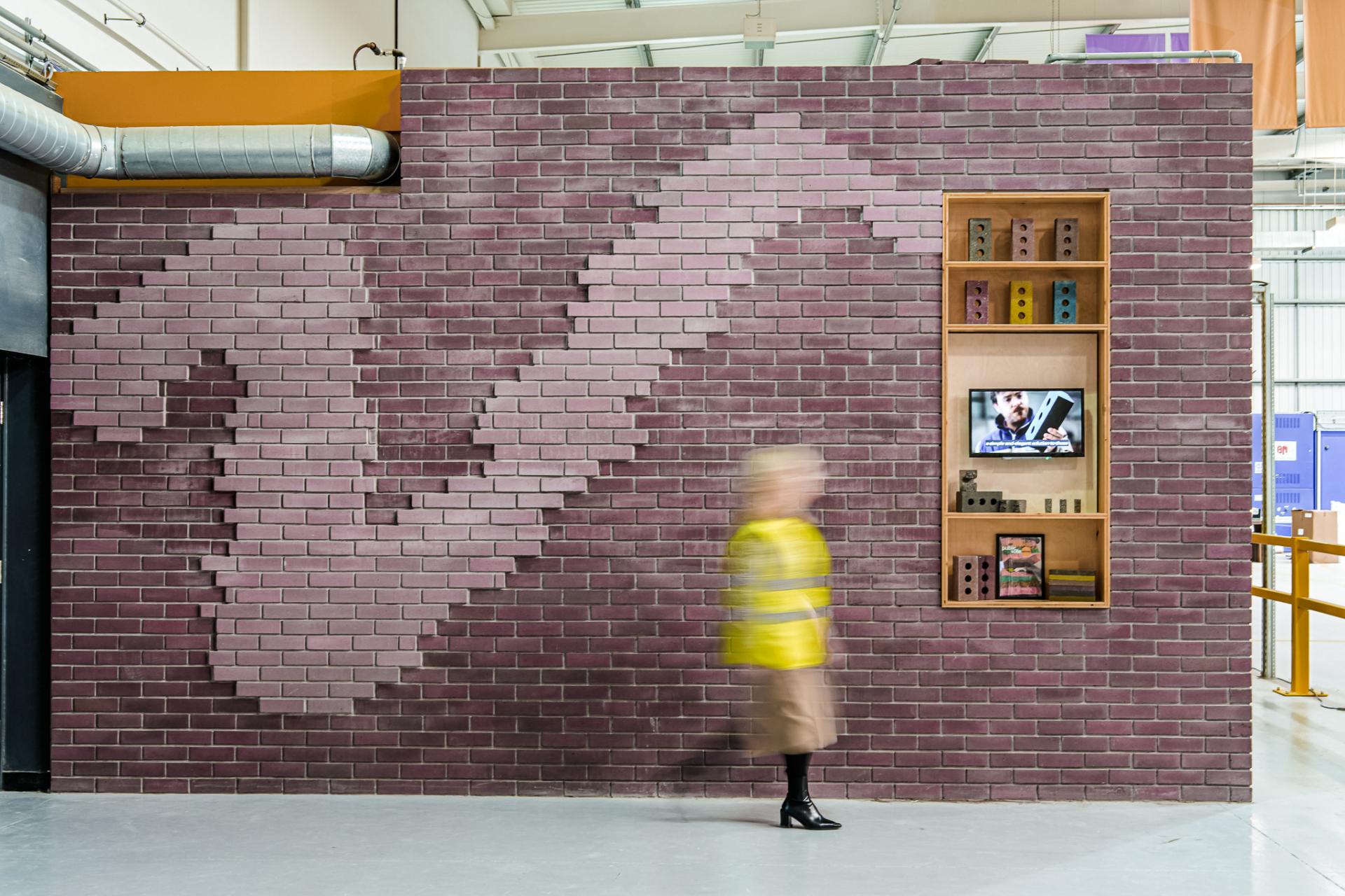 A woman in a yellow jacket walks past a wall showcasing innovative un-fired, ultra low carbon bricks.