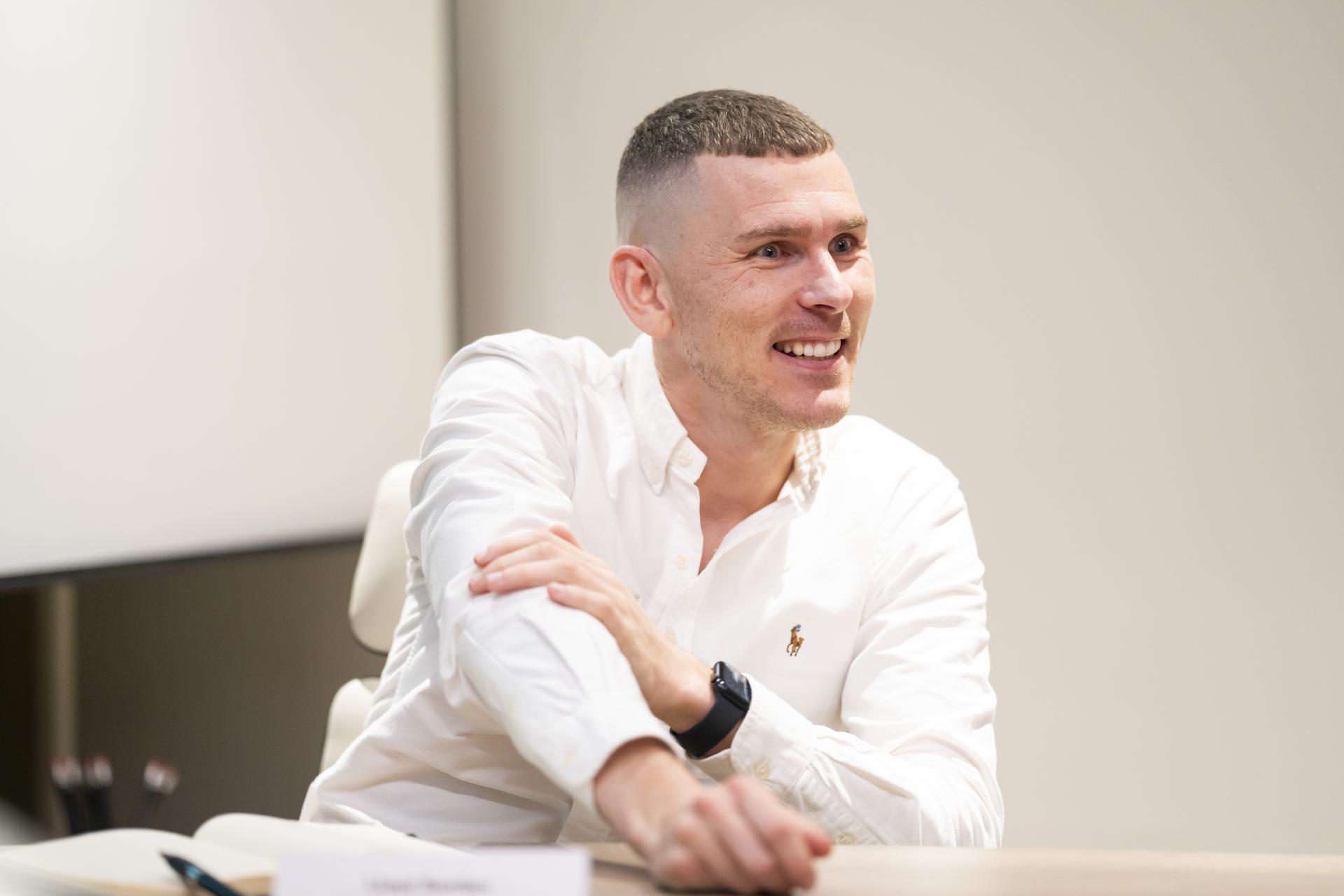 A smiling man in a white shirt discusses the role of technology in sustainable design at a meeting.
