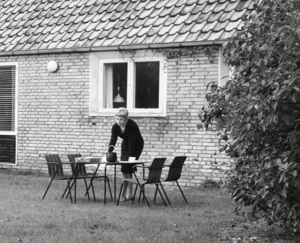 Woman preparing a table outdoors near a brick house, showcasing Danish design influence and mid-century aesthetics.