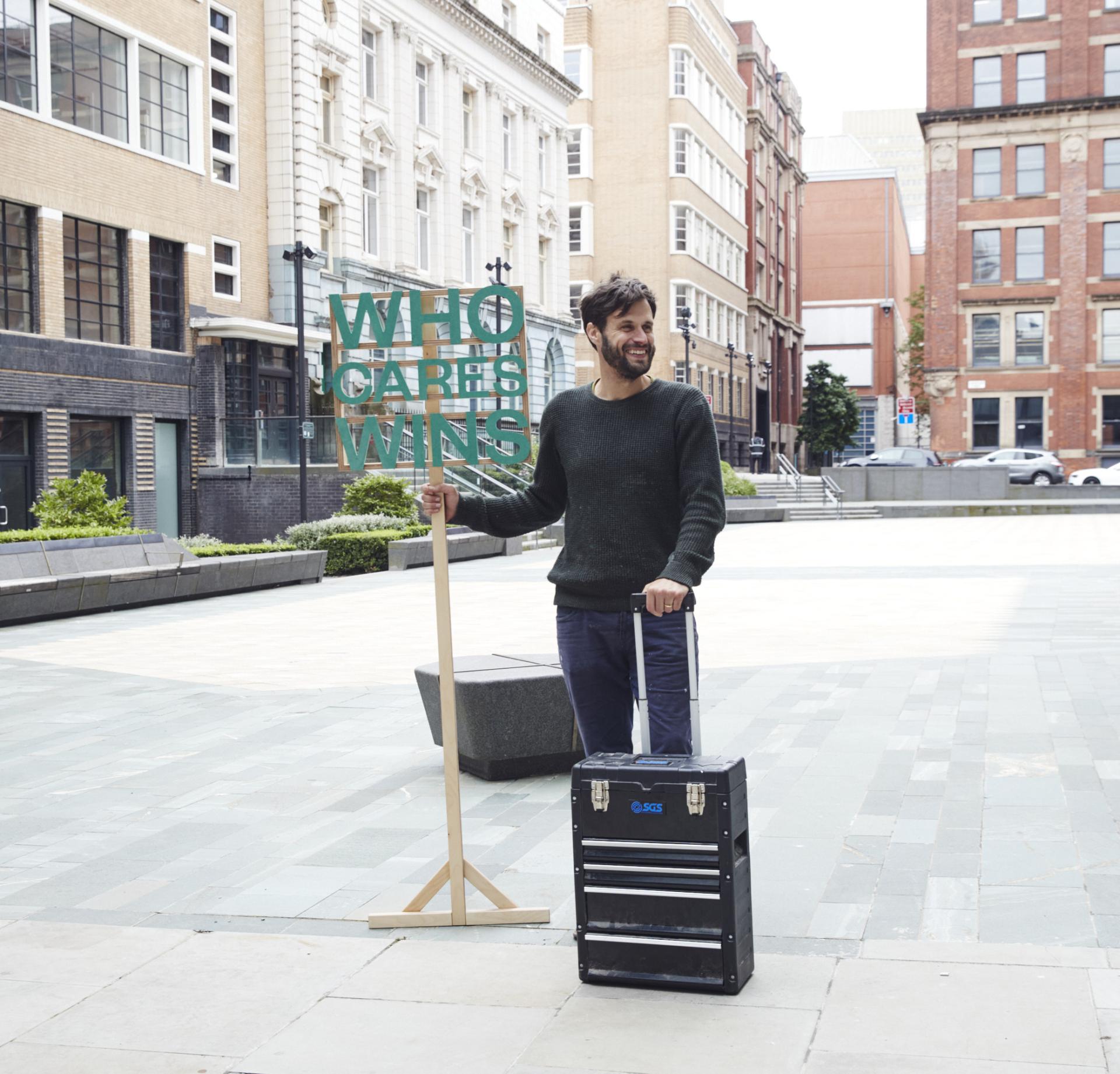 Man holding an uplifting "WHO CARES WINS" sign in Manchester's NOMA neighbourhood, representing community spirit and collaboration.