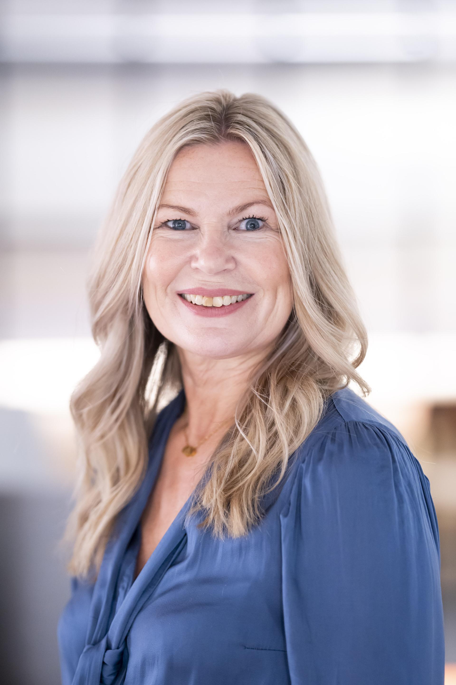 Smiling woman with long blonde hair wears a blue blouse, representing workplace engagement in a modern office setting.
