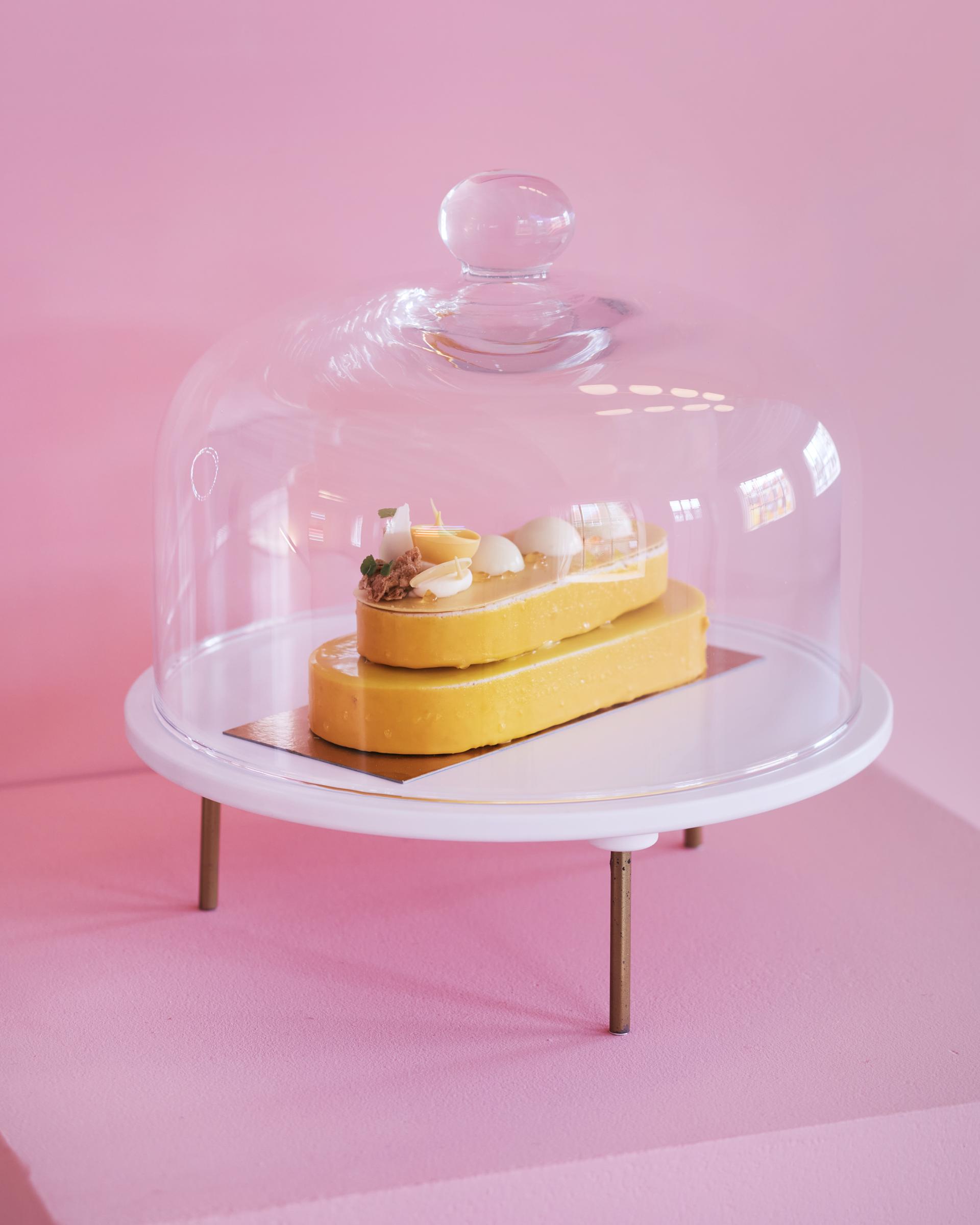 A vibrant yellow dessert under a glass dome, showcasing playful patisserie-style design against a pink background.