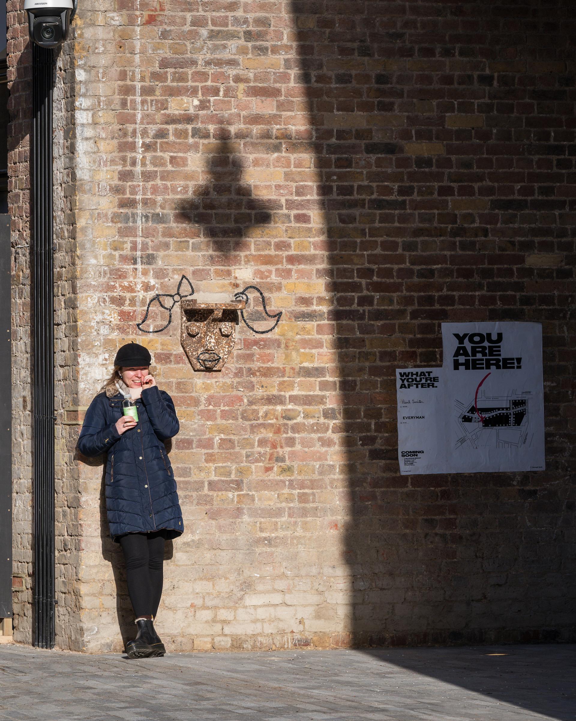Young woman enjoying a drink while leaning against a brick wall decorated with art and directional signage at Borough Yards, London.