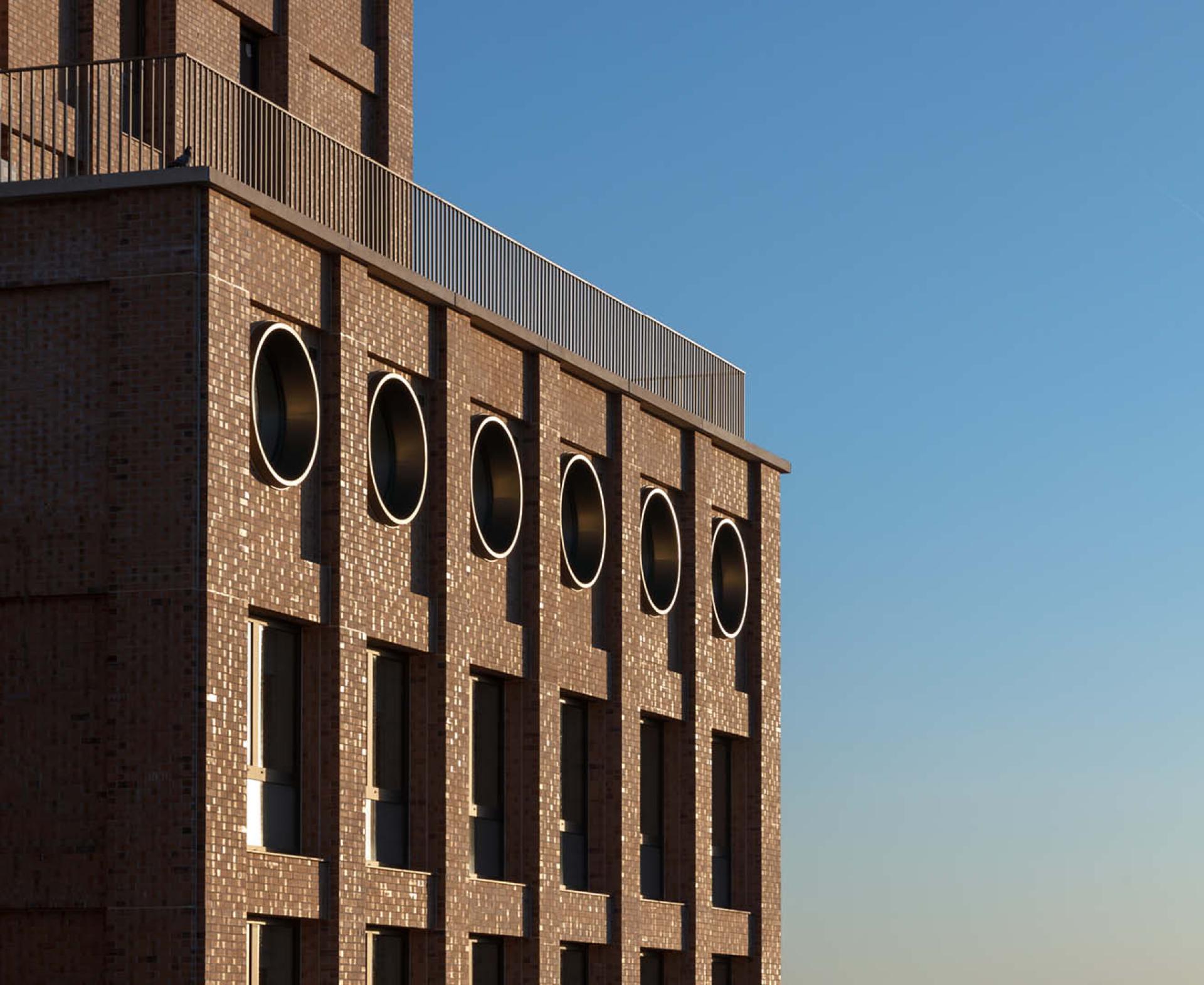 Modern co-living building with circular windows and textured brick facade, highlighting innovative architectural design amidst a clear blue sky.
