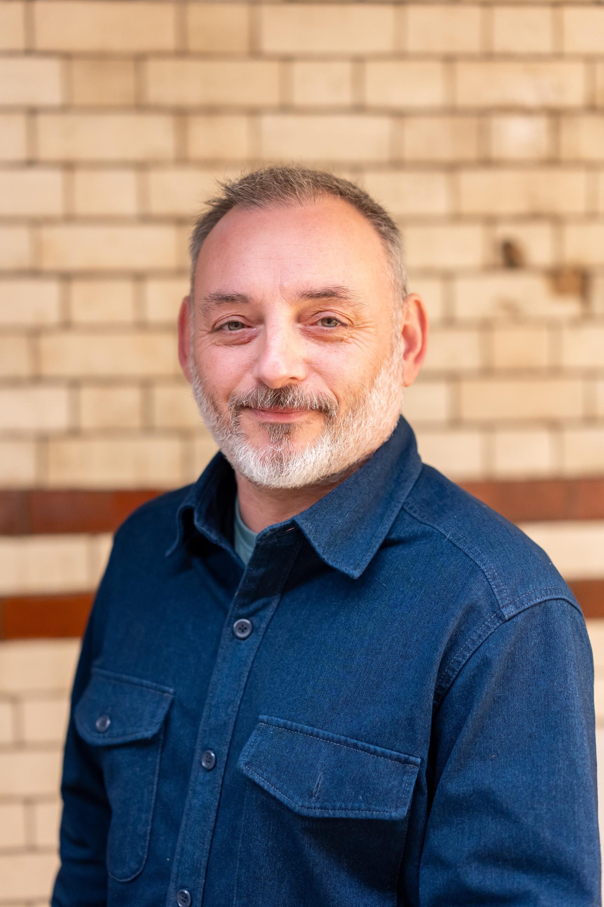 Man in a denim shirt smiles confidently in front of a brick wall, embodying a positive cultural approach to EDI.