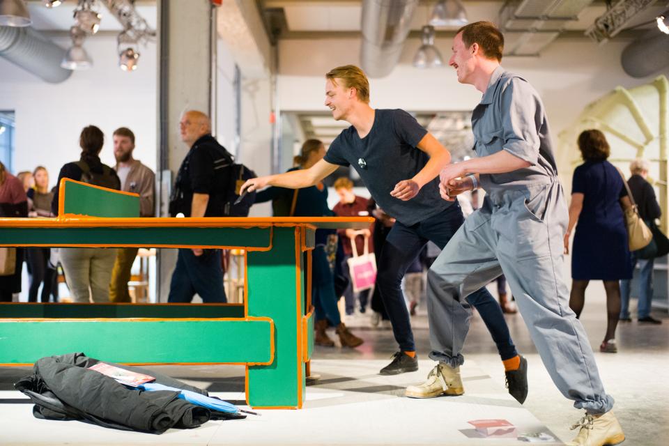 Two men engaged in playful interaction with a colorful table installation at Dutch Design Week, surrounded by visitors.