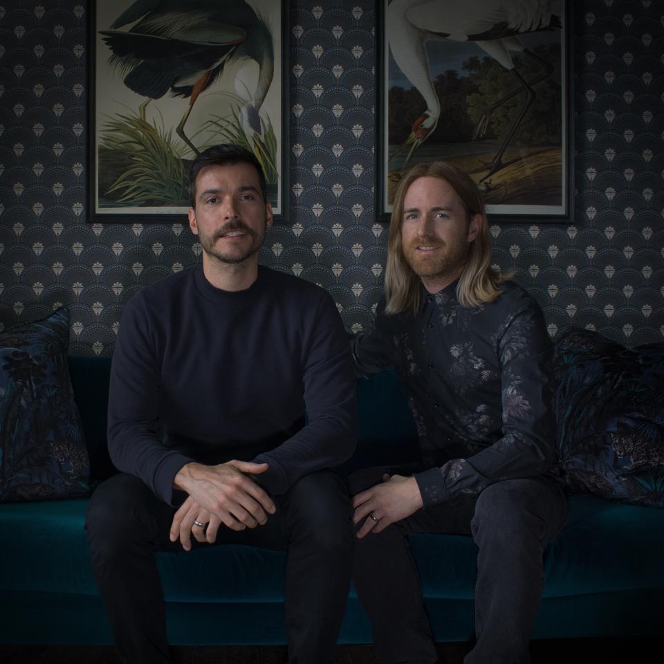 Design duo Divine Savages seated on a teal couch, showcasing elegant wallpapers and luxury interiors inspiration.