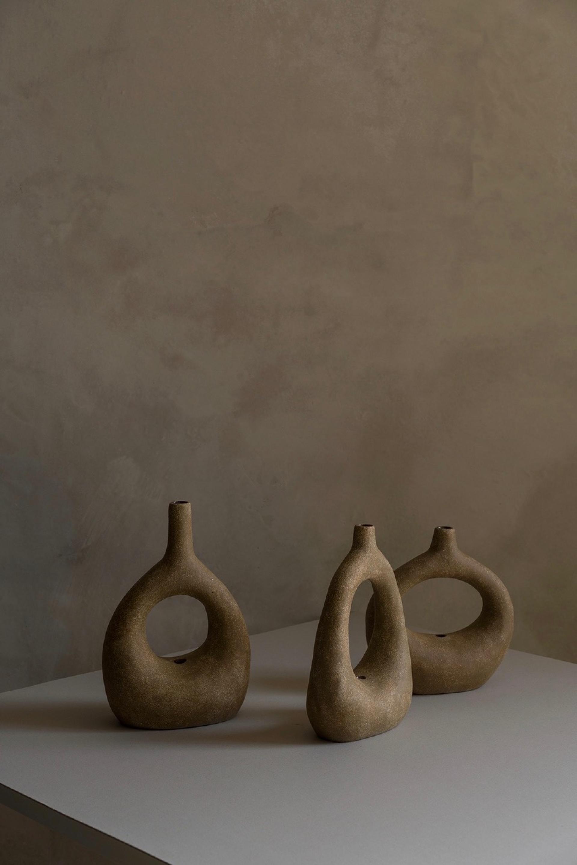 Three uniquely shaped ceramic vases in earthy tones, displayed on a minimalistic table against a textured backdrop.