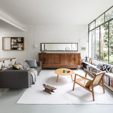 Renovated mid-century apartment interior with a cozy living area, wooden furniture, and large windows overlooking lush gardens.