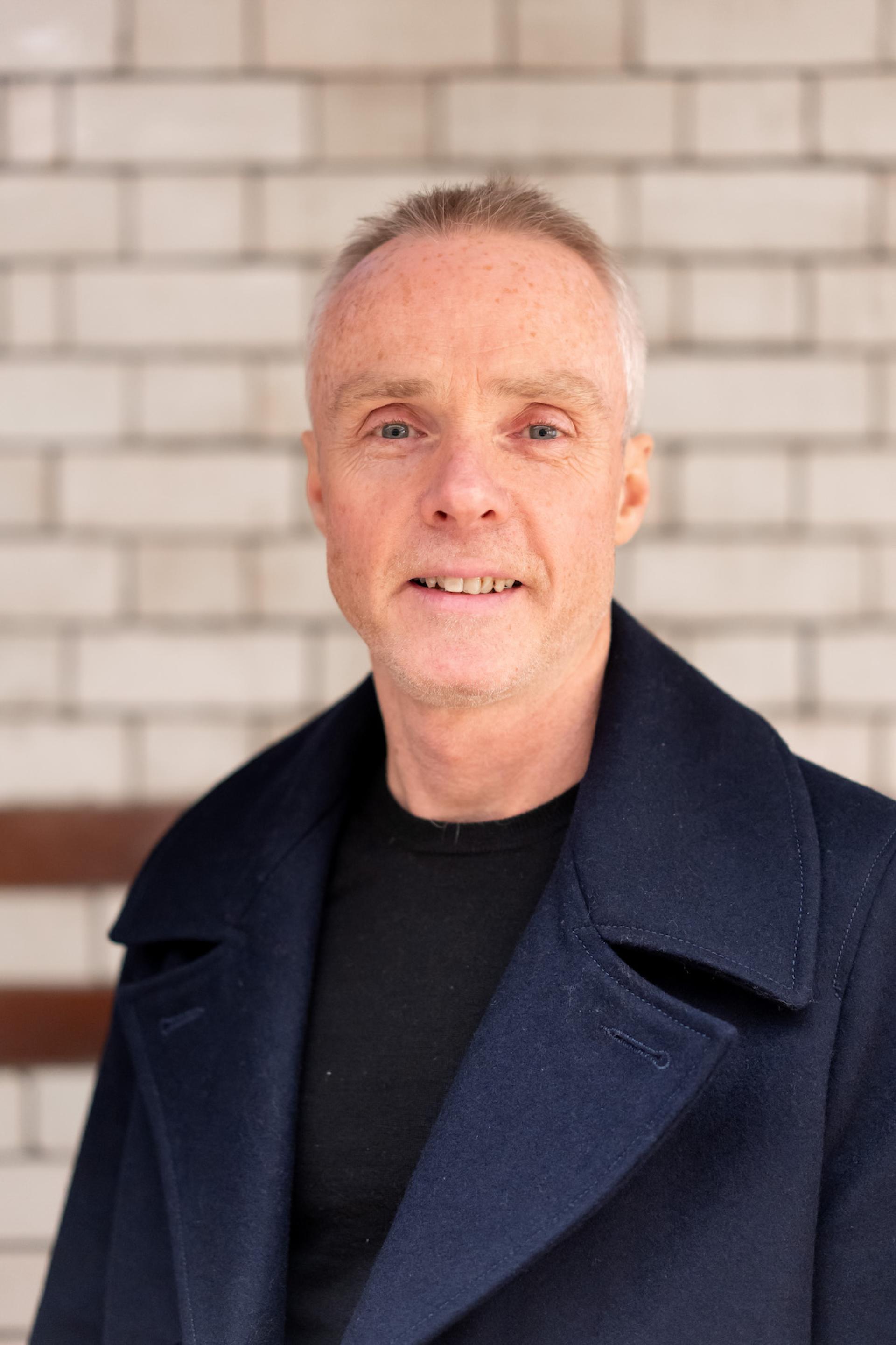 A man in a navy coat smiles confidently, discussing sustainability and value engineering at a roundtable seminar.