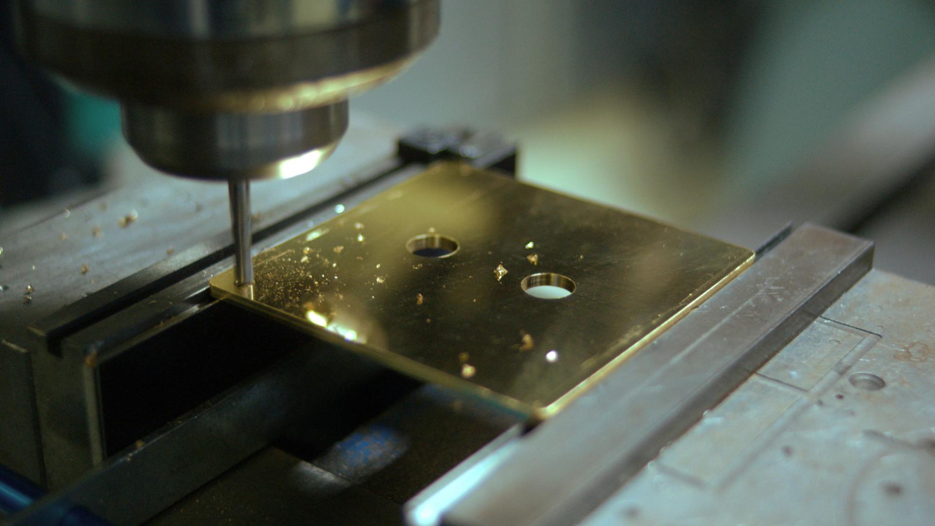 Metal machining process showing a gold-colored plate being drilled, illustrating Lutron's manufacturing quality for HomeWorks products.